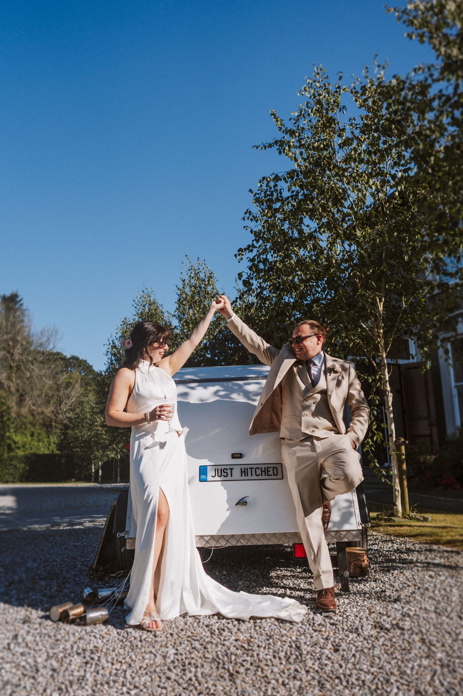 A couple dances and poses playfully together by a white outdoor hot tub on a gravel patio in sunlit garden setting.