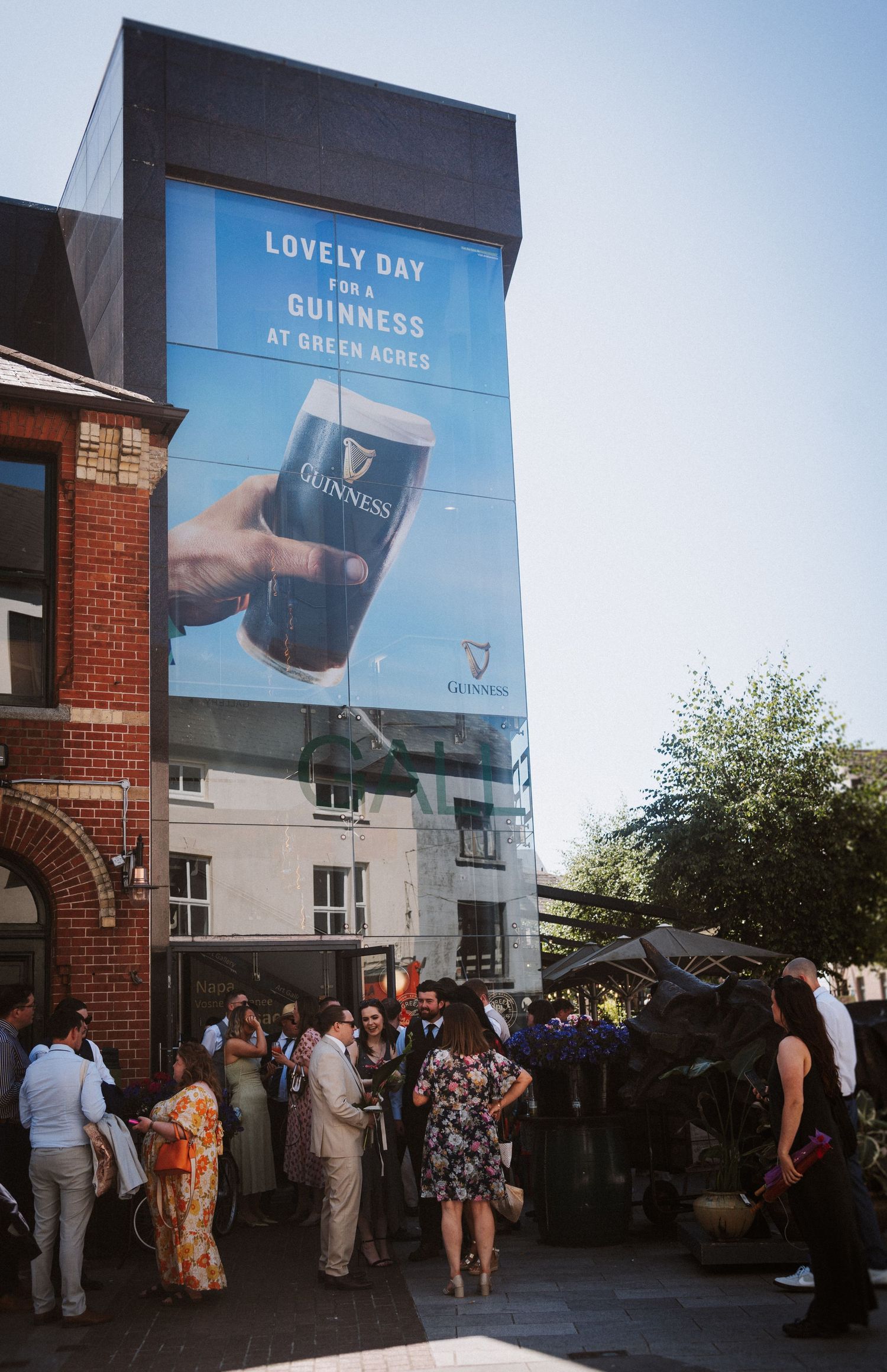 A group of people gather outside a brick building with Guinness advertising banners displayed in the summer evening light.