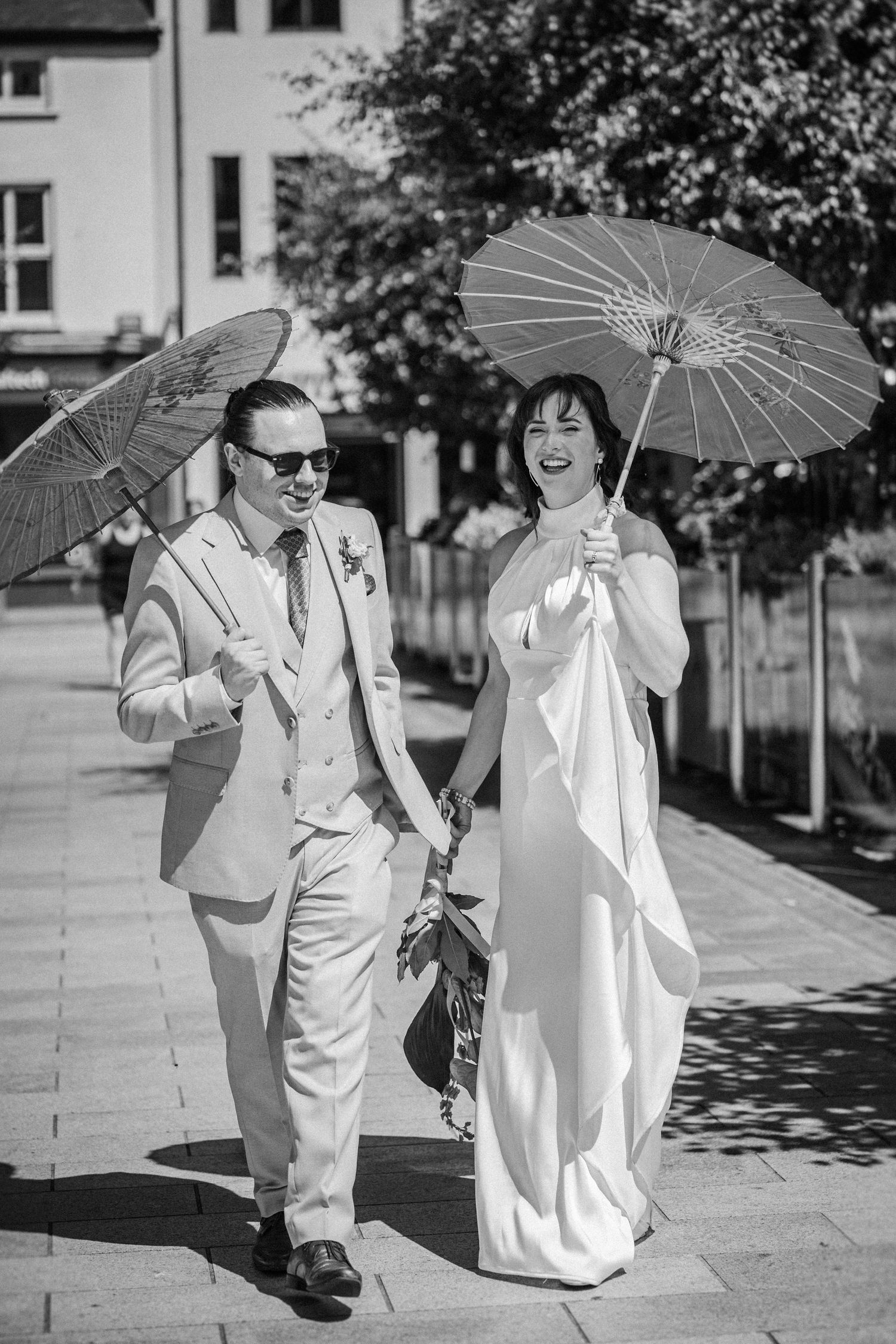 A couple in vintage attire walk playfully together carrying parasols on a sunny sidewalk.