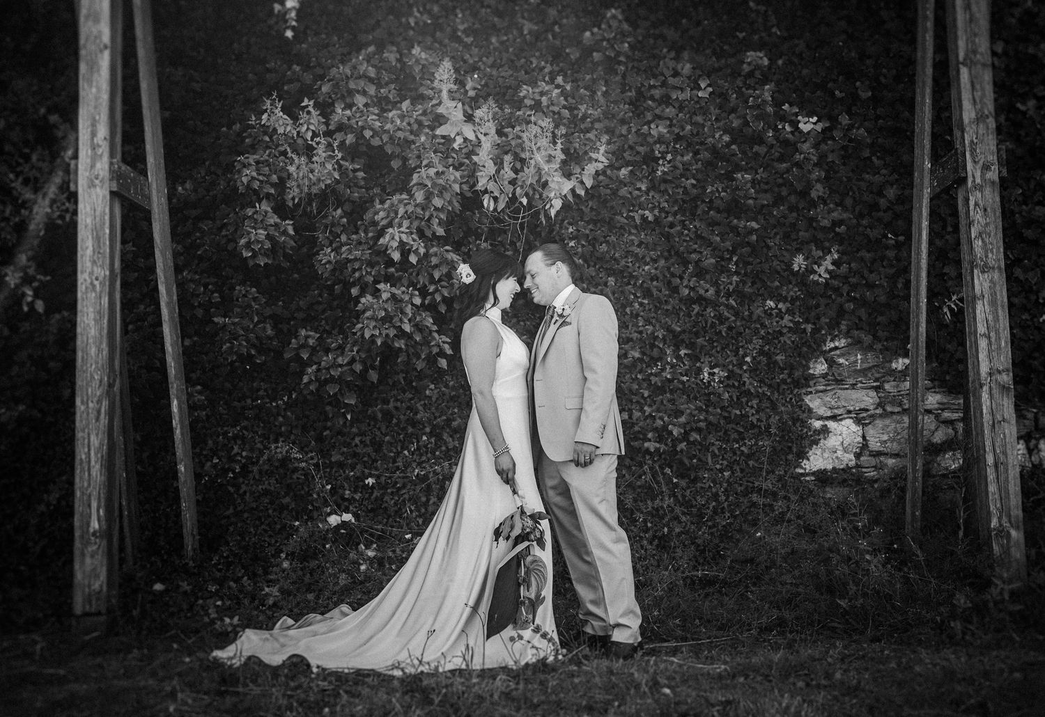 Black and white photo showing a romantic couple holding hands while walking through a grove of tall trees at dusk.