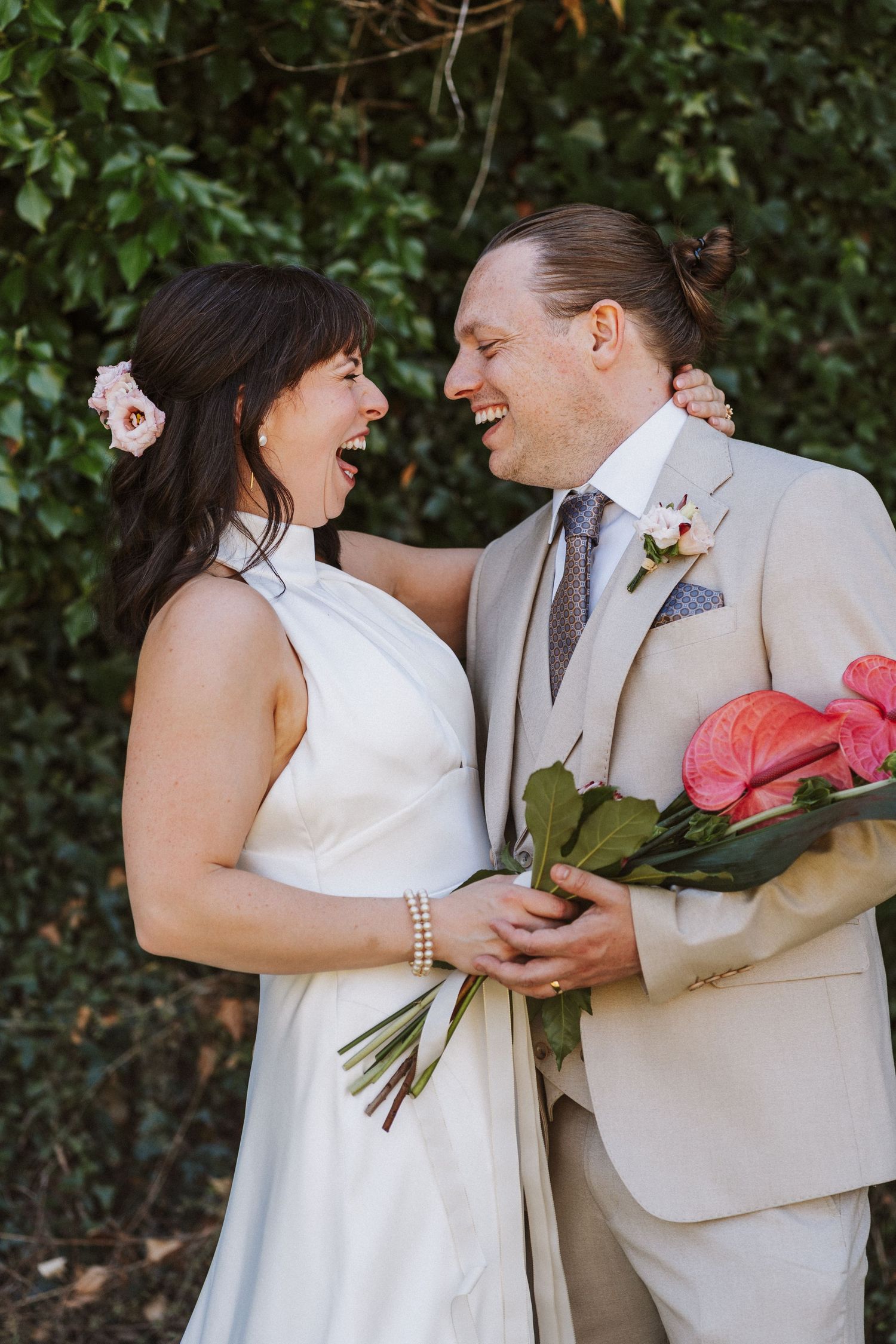 A couple laughing and embracing during an outdoor wedding photoshoot with greenery in the background.