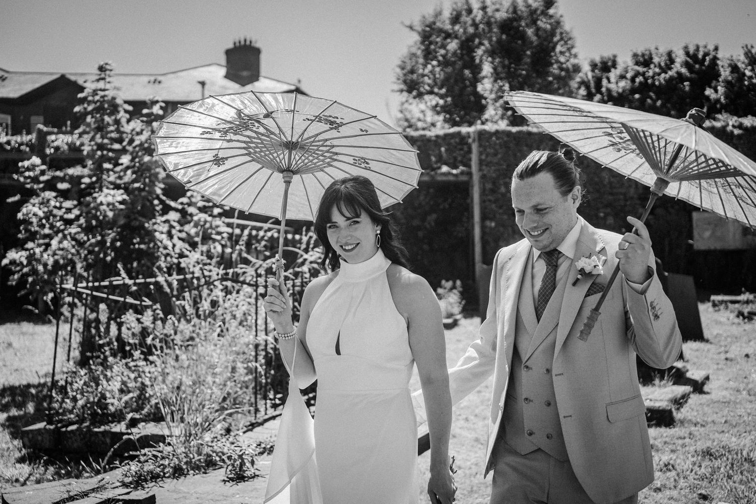 A black and white photo of a couple walking with parasols on a sunny garden path.