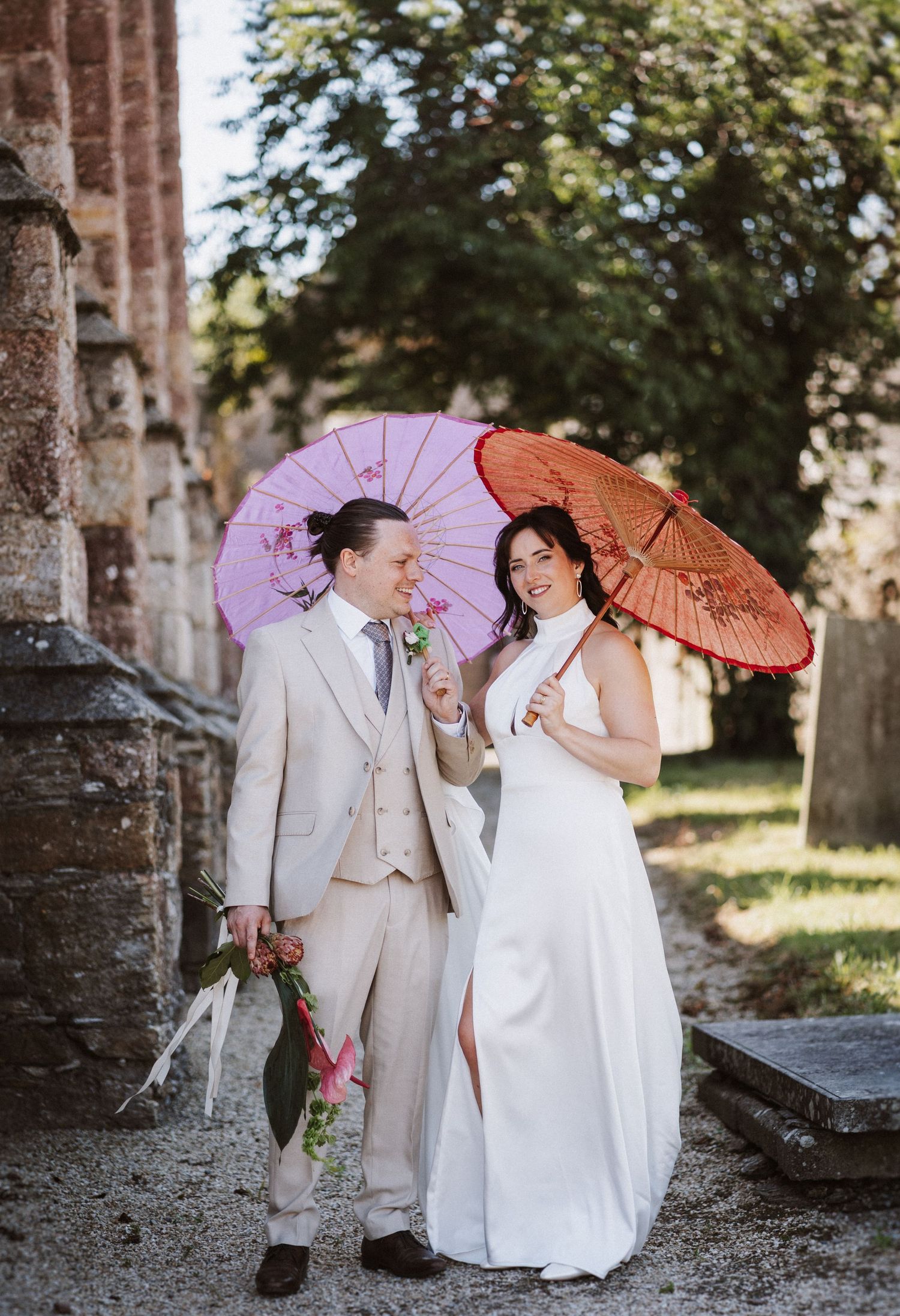 A couple shares a playful moment with pink and orange paper parasols while posing together by stone ruins at sunset.