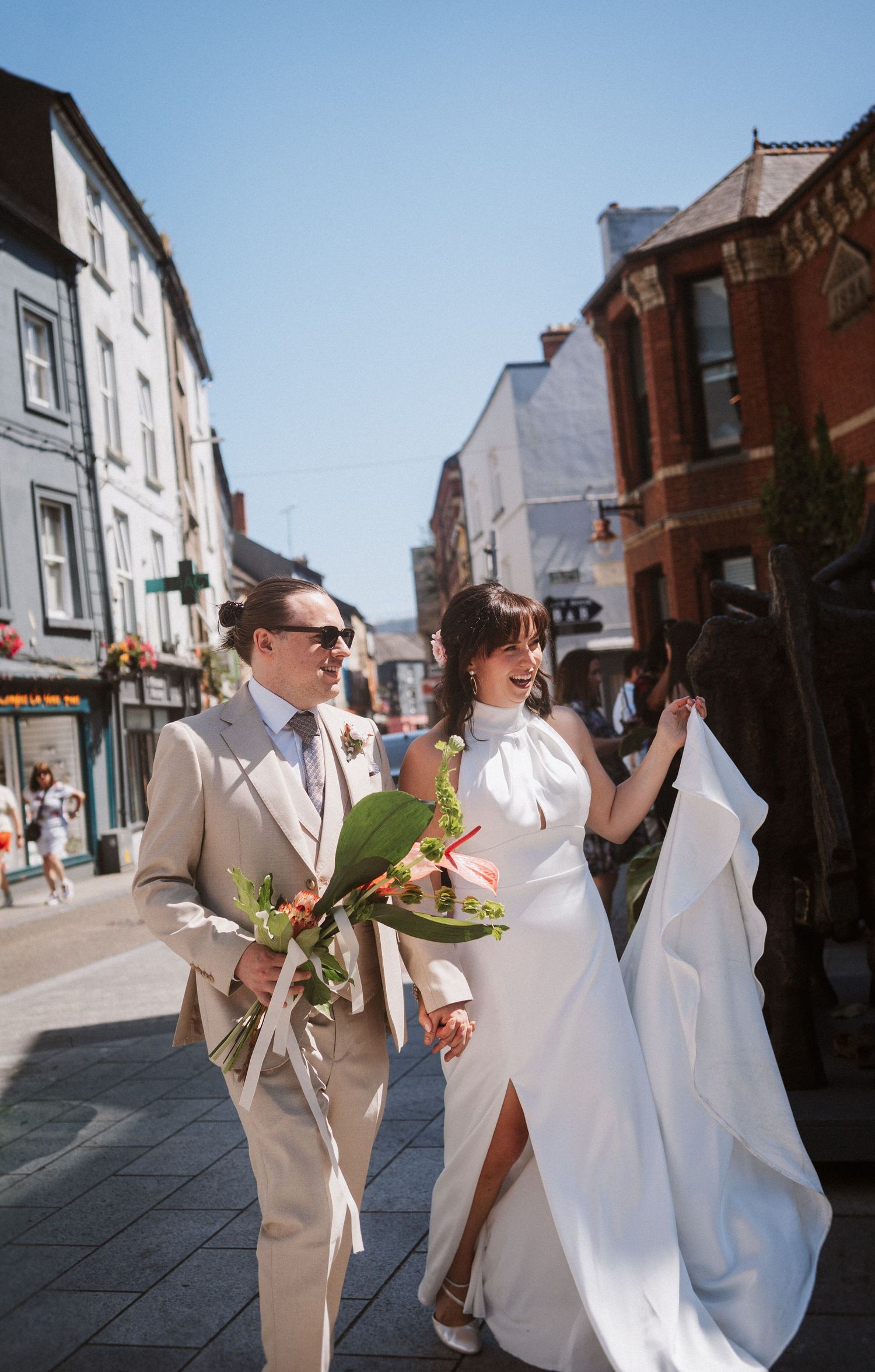 A couple walking down a sunny street while holding hands and drinking wine from plastic cups.