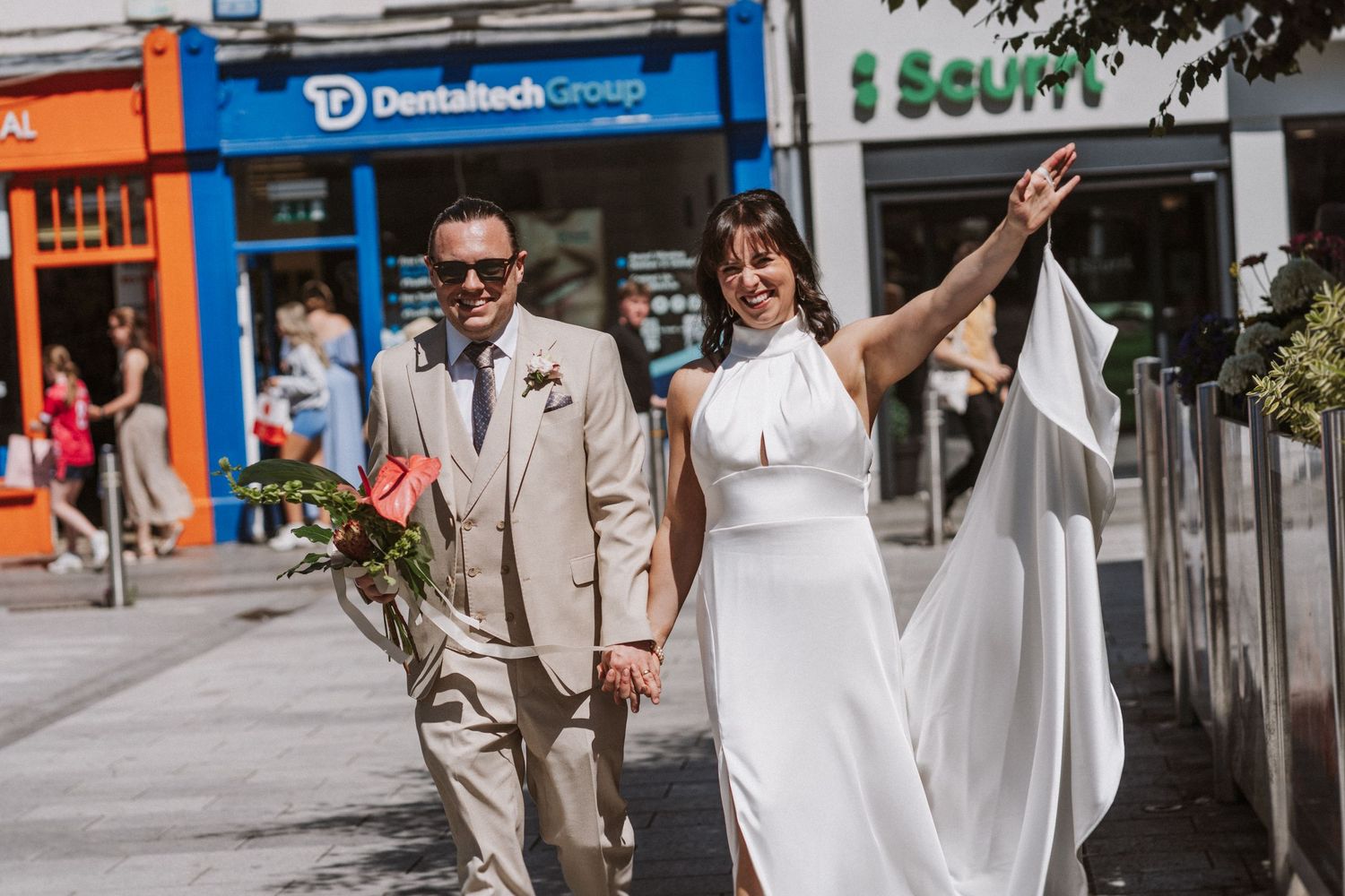 Happy couple walks down urban sidewalk holding hands while wearing matching beige and white outfits on their wedding day.