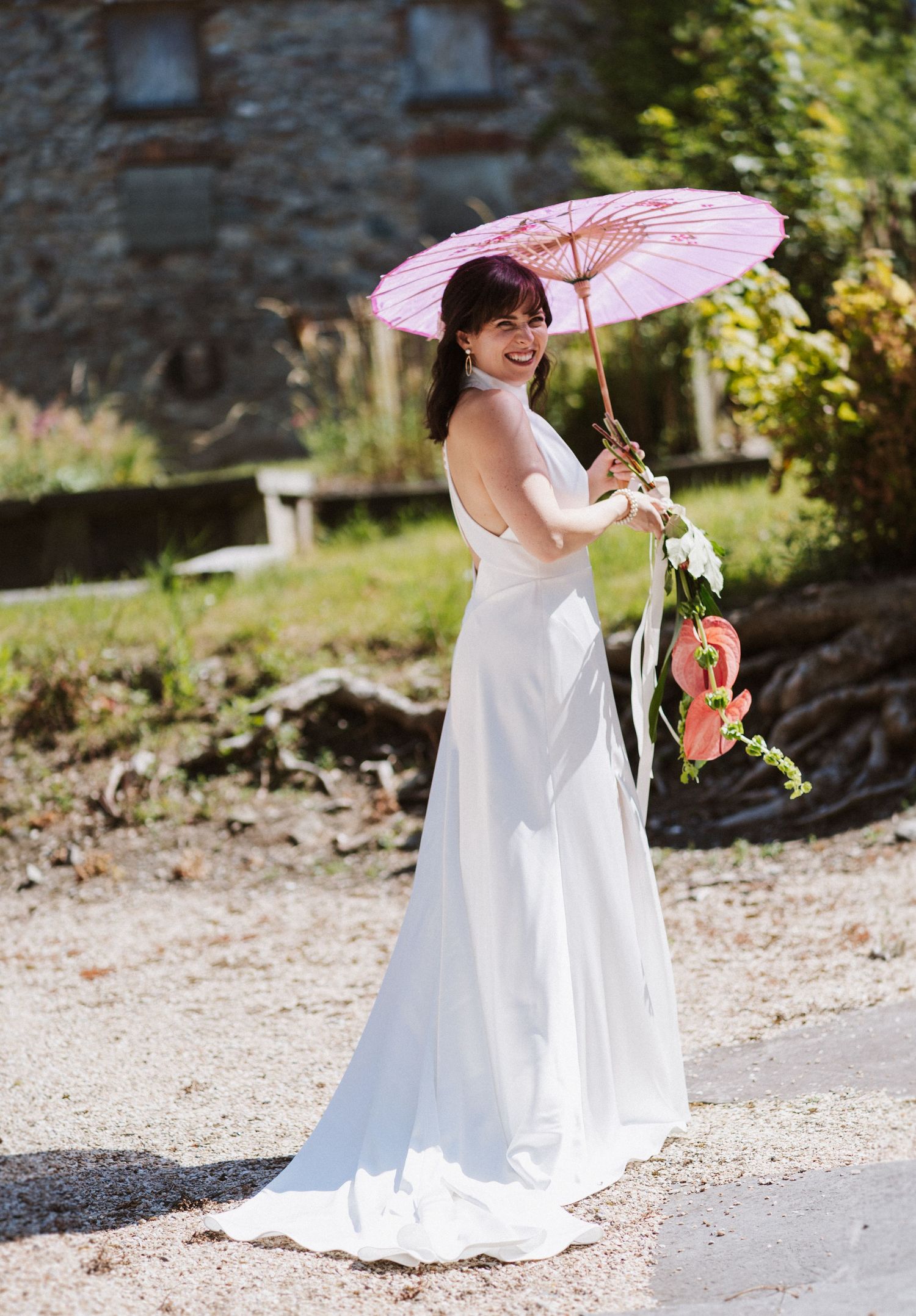 A person in a white wedding dress holding a pink parasol poses elegantly on a gravel path on a sunny day.