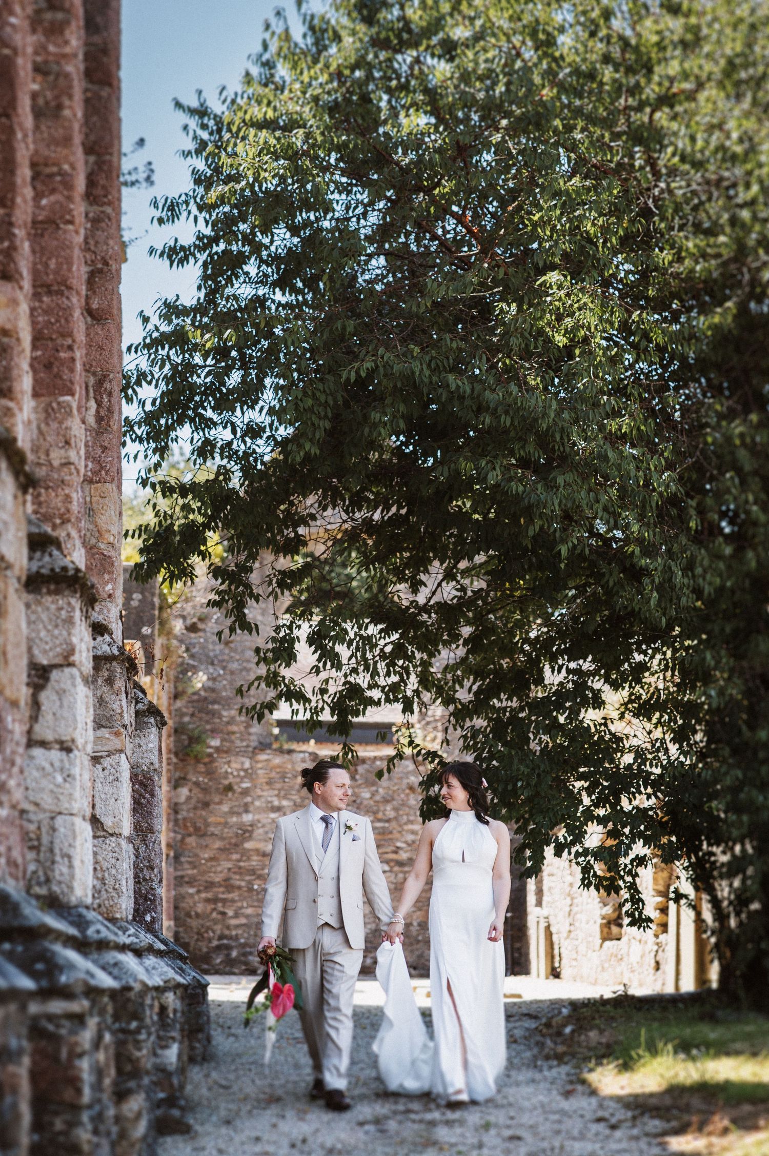 Wedding couple walking through an archway of historic stone ruins surrounded by green trees on a sunny day.