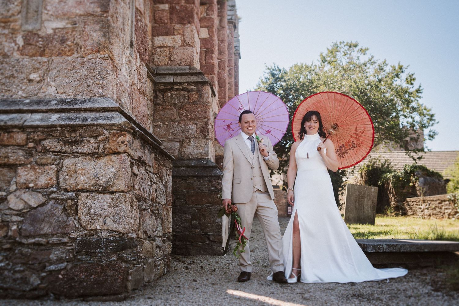 Wedding couple embraces in front of rustic stone church wall during outdoor countryside celebration.