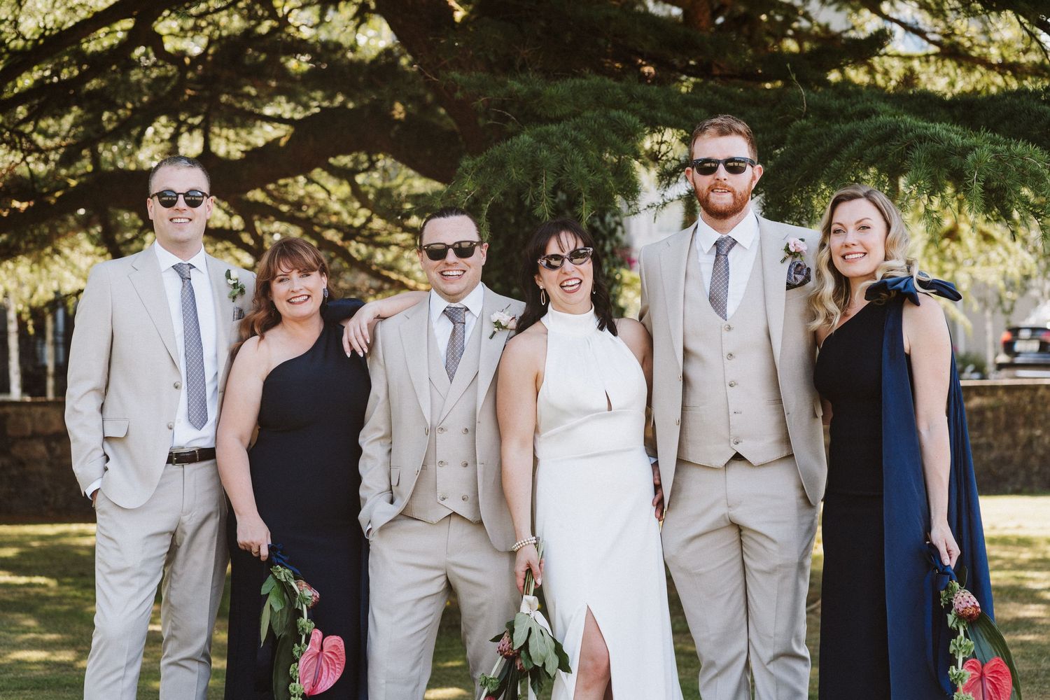 Wedding party in light beige suits and formal attire poses together under large autumn trees for outdoor photos.