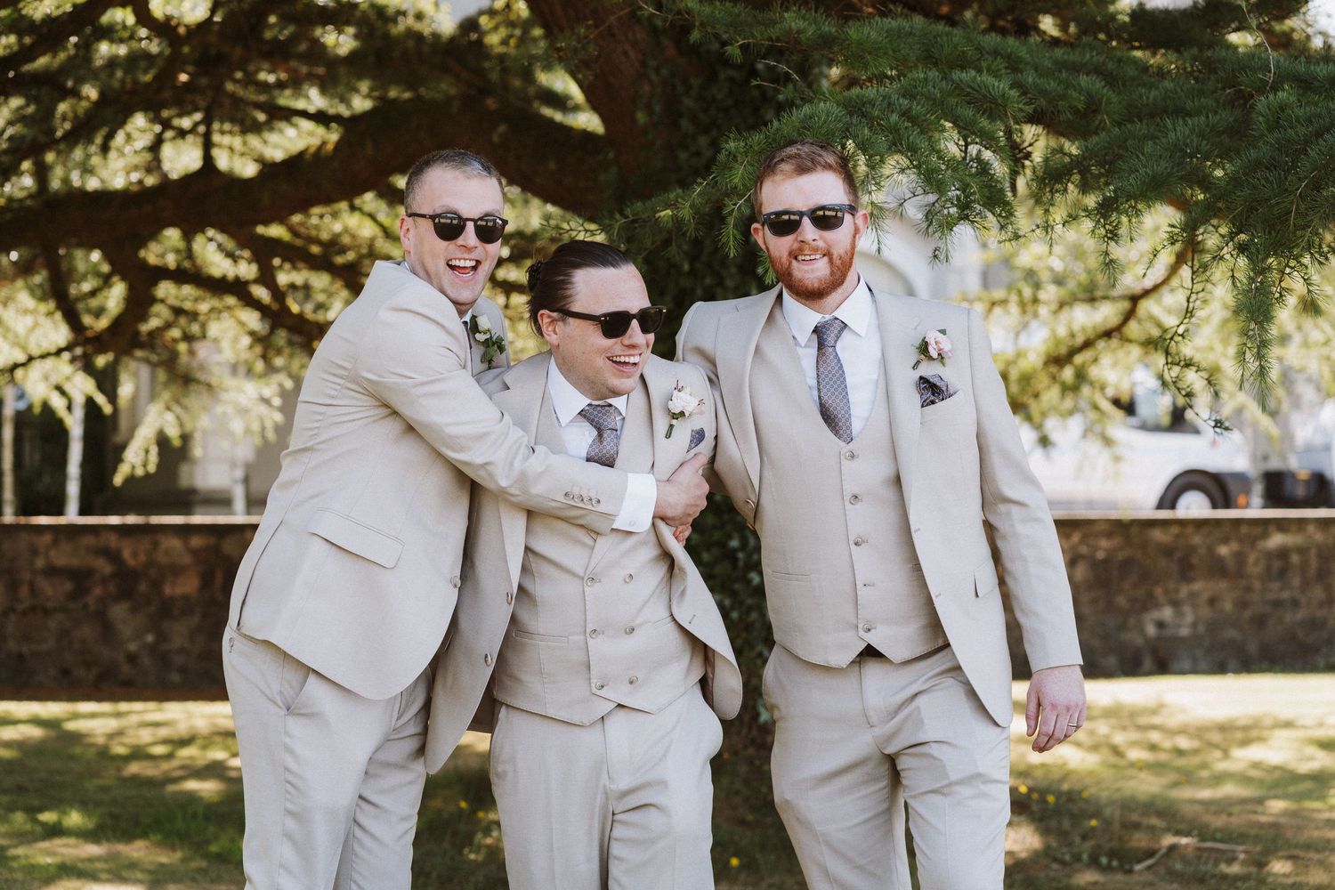 Groomsmen in light beige suits walk together outdoors on a sunny day.