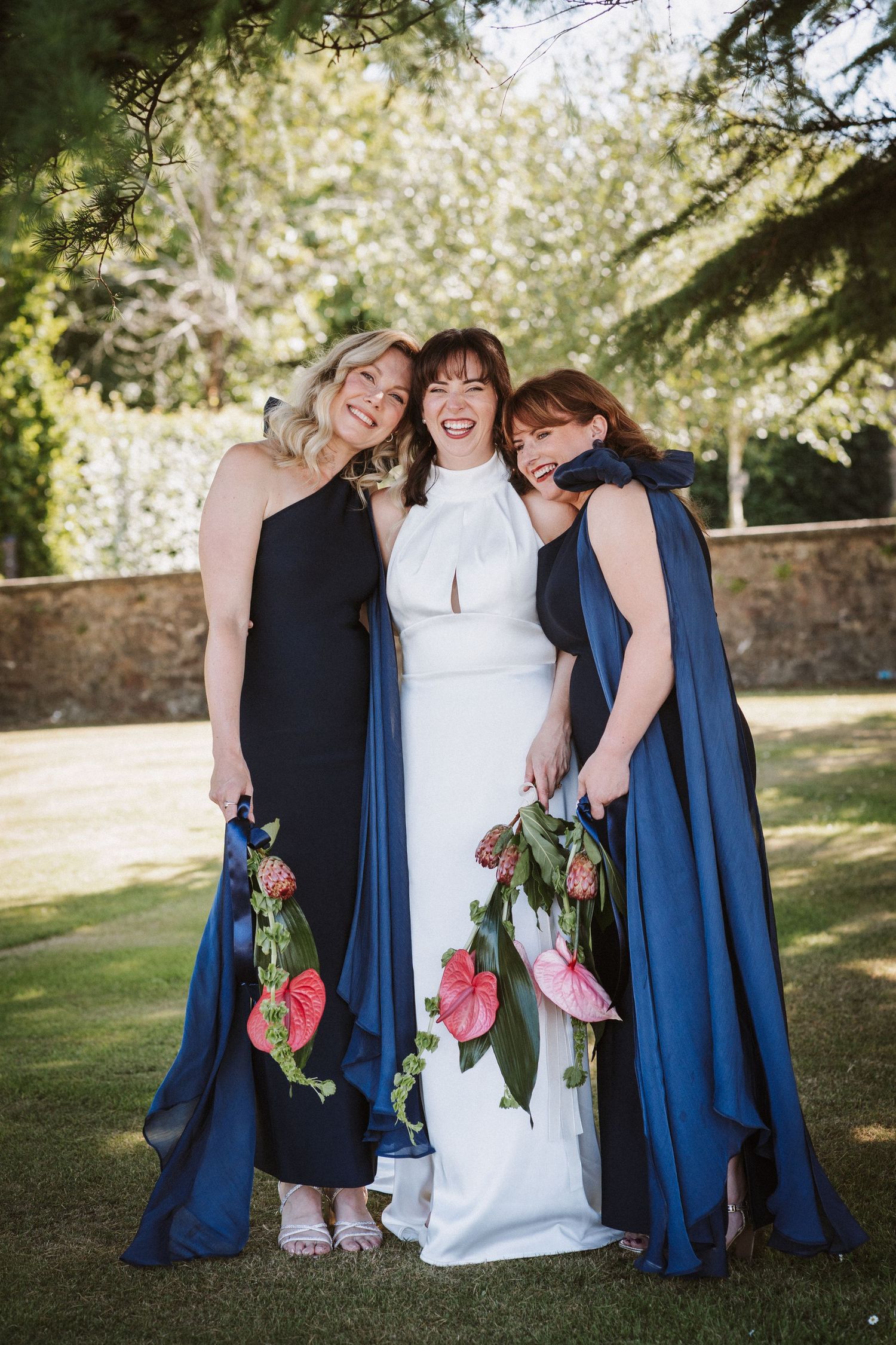 Bridesmaids in navy blue dresses share joyful laughs with bride in white dress while posing outdoors in springtime setting.