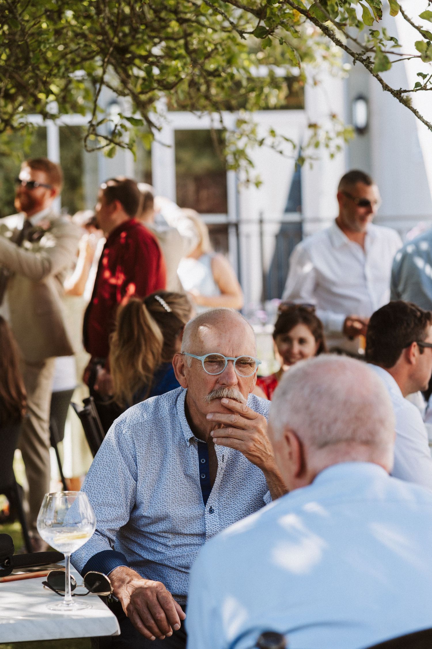 Seniors enjoy conversation at an outdoor restaurant table with wine glasses under dappled sunlight.