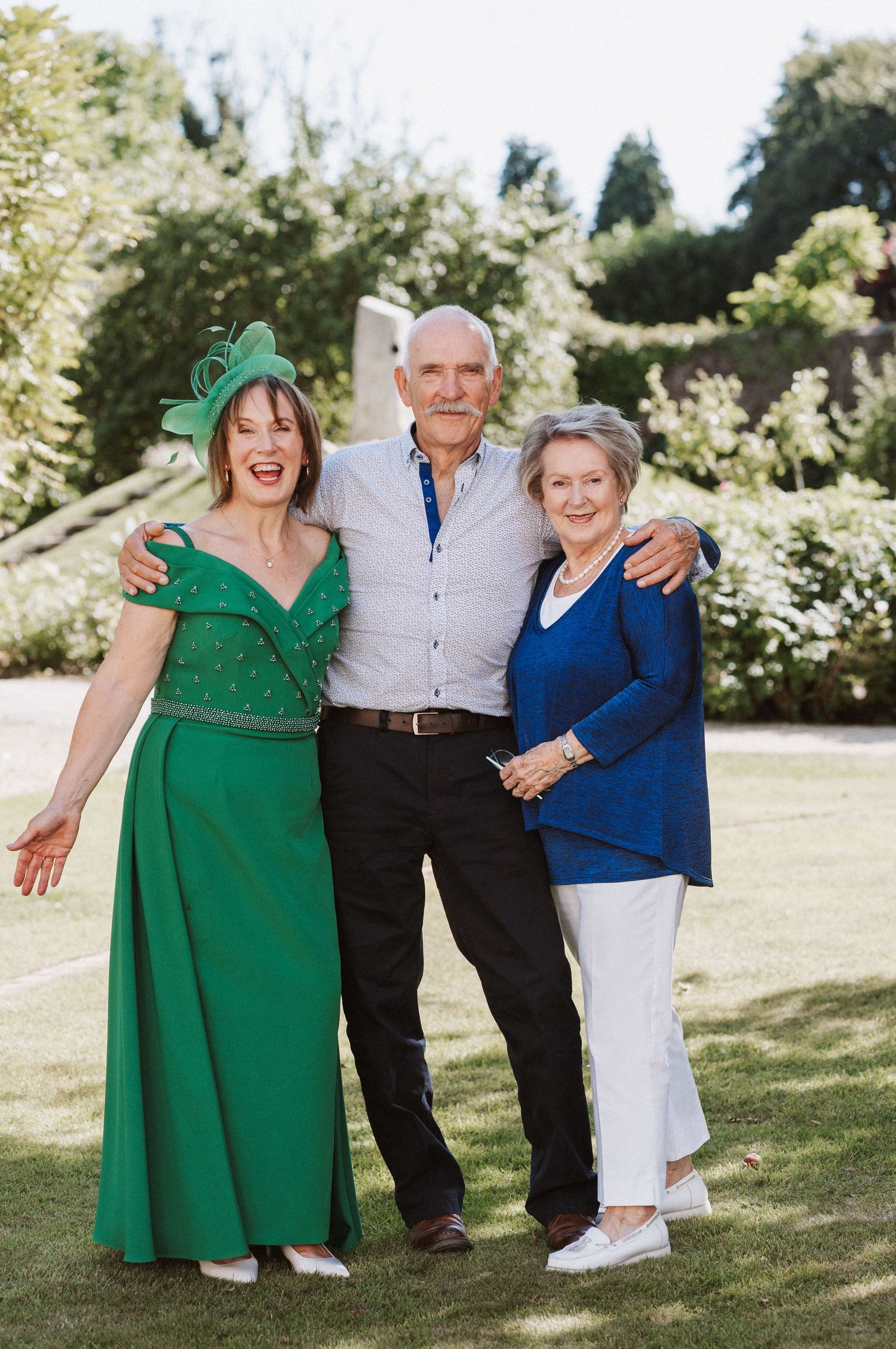 Three people standing together outdoors on a sunny lawn, with one wearing an emerald green dress and another in navy blue.