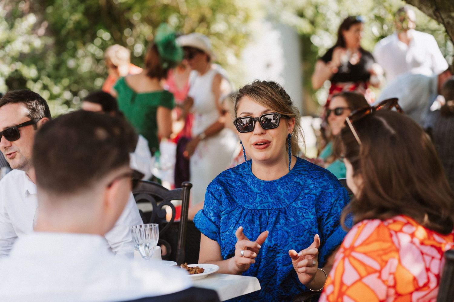 A crowd of people wearing sunglasses sits outdoors at tables enjoying a casual gathering in natural lighting.
