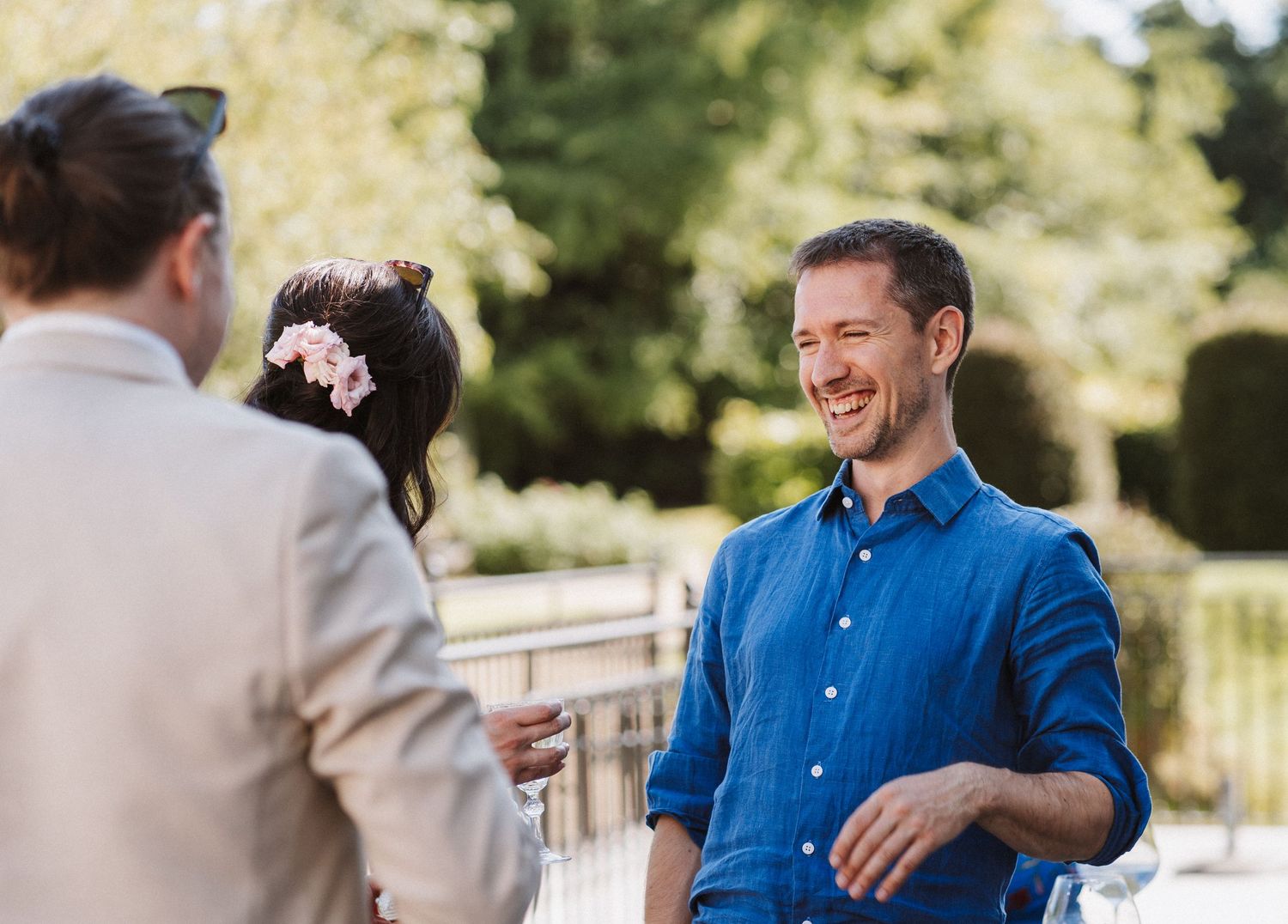 Wedding party sharing laughs and conversation on outdoor wooden deck during daytime celebration.