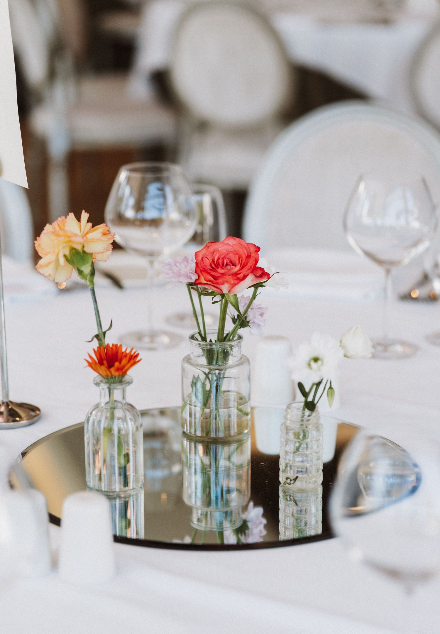 Simple glass jars with fresh flowers serve as rustic centerpieces on a white tablecloth at a wedding reception.