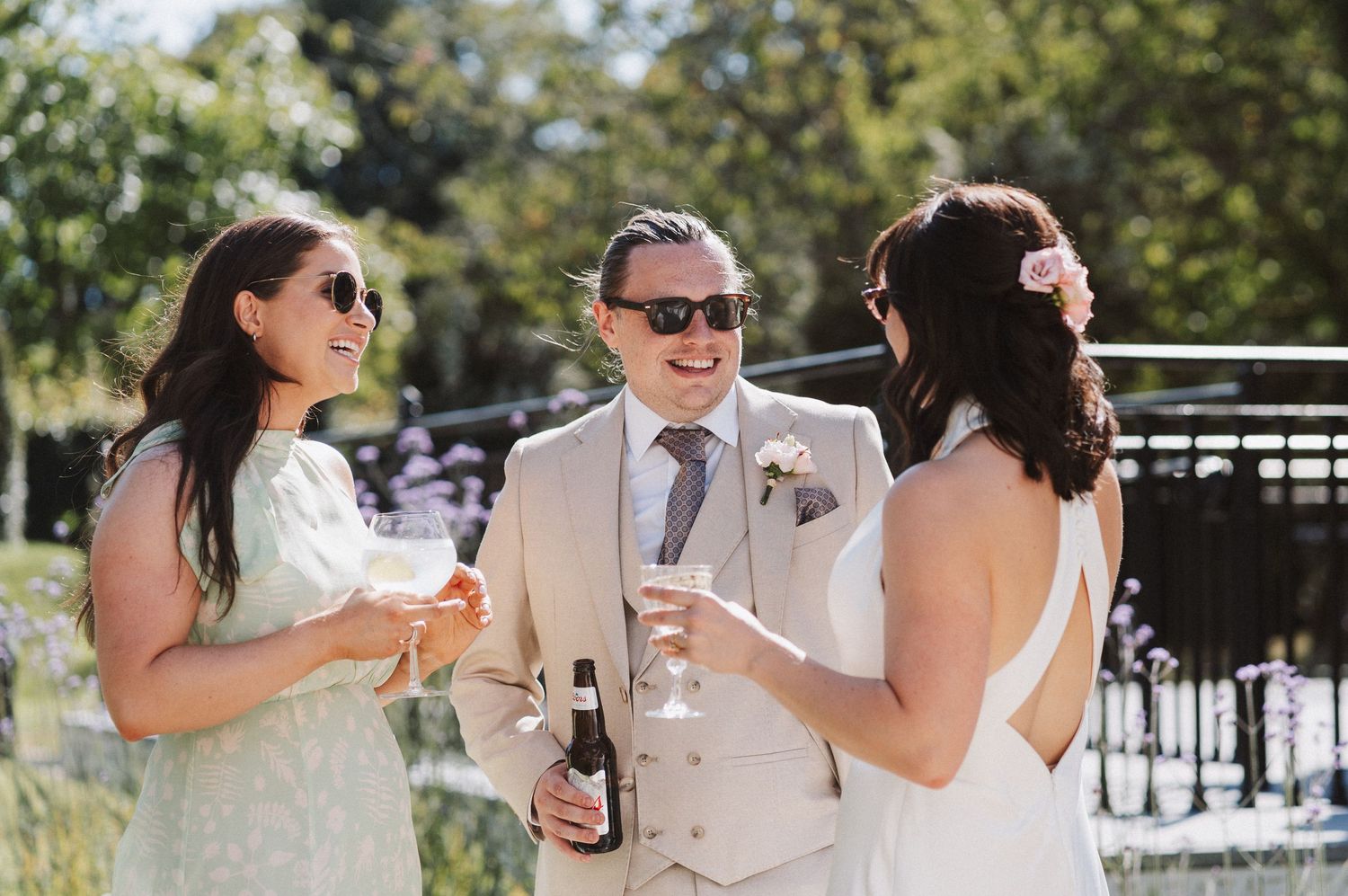Three people wearing elegant attire enjoy drinks while conversing outdoors at a wedding celebration on a sunny day.