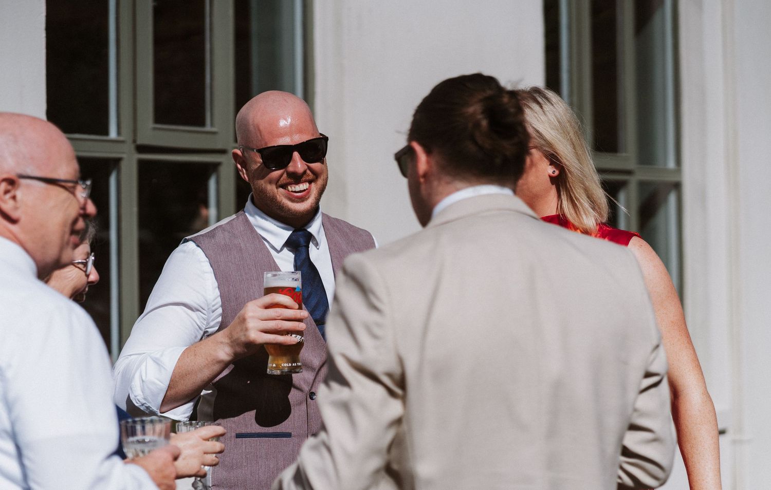 Well-dressed guests in sunglasses and light-colored attire enjoy drinks at an outdoor social gathering.