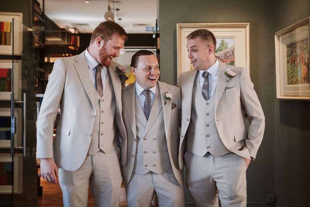 A group of groomsmen in matching light gray suits share a candid laughing moment indoors.