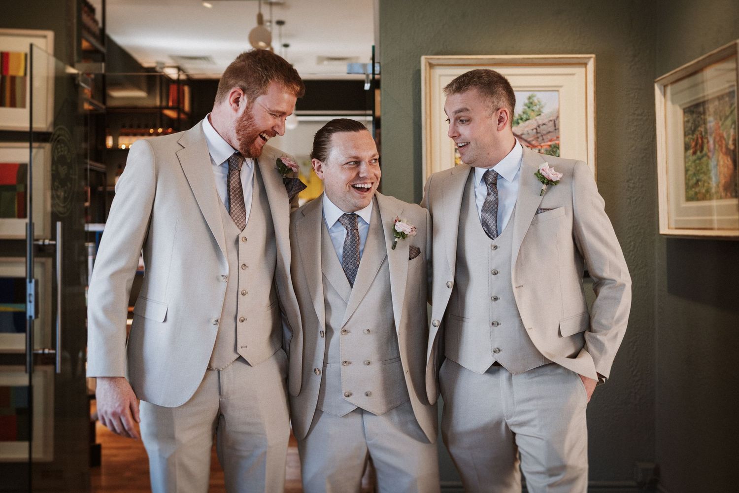 A group of groomsmen in matching light gray suits share a candid laughing moment indoors.