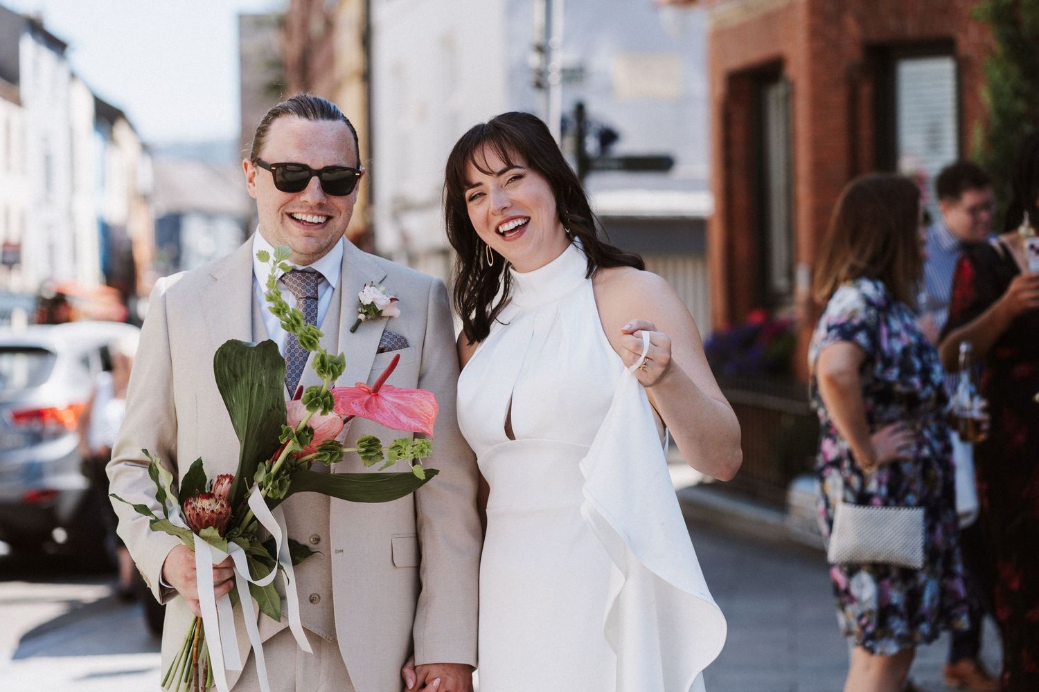 A joyful couple celebrates their wedding day on a sunny urban street while holding a floral bouquet.