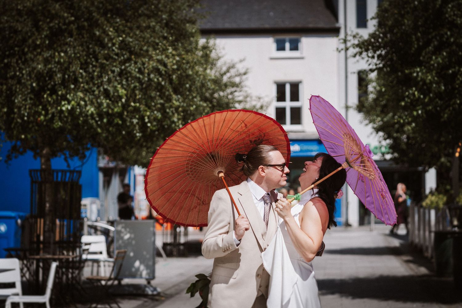 A group of wedding guests in traditional dress hold colorful parasols outdoors in front of a building.