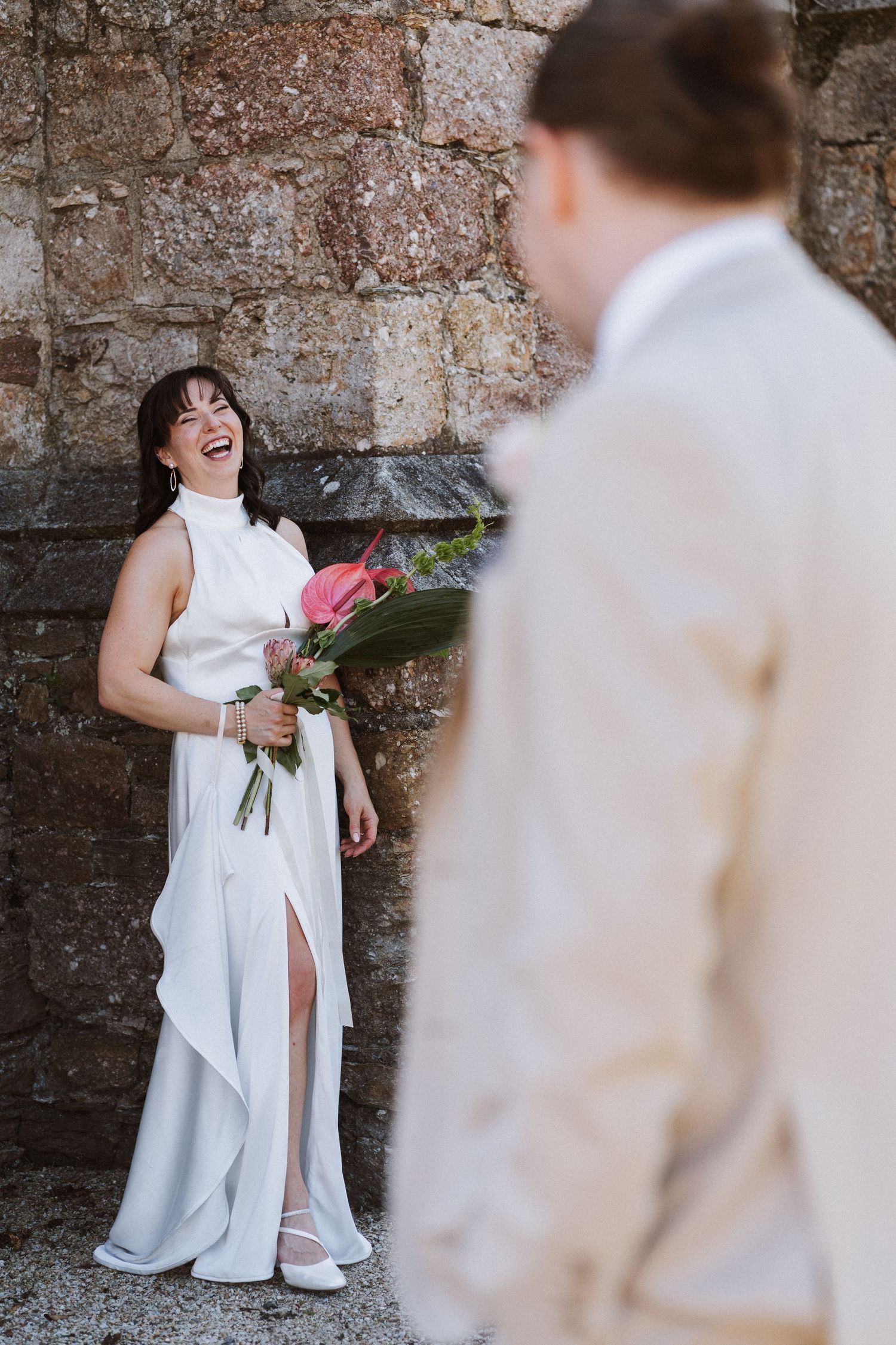 A joyful wedding moment captured against a stone wall as a bride in a white dress holds a bouquet, laughing with her partner.