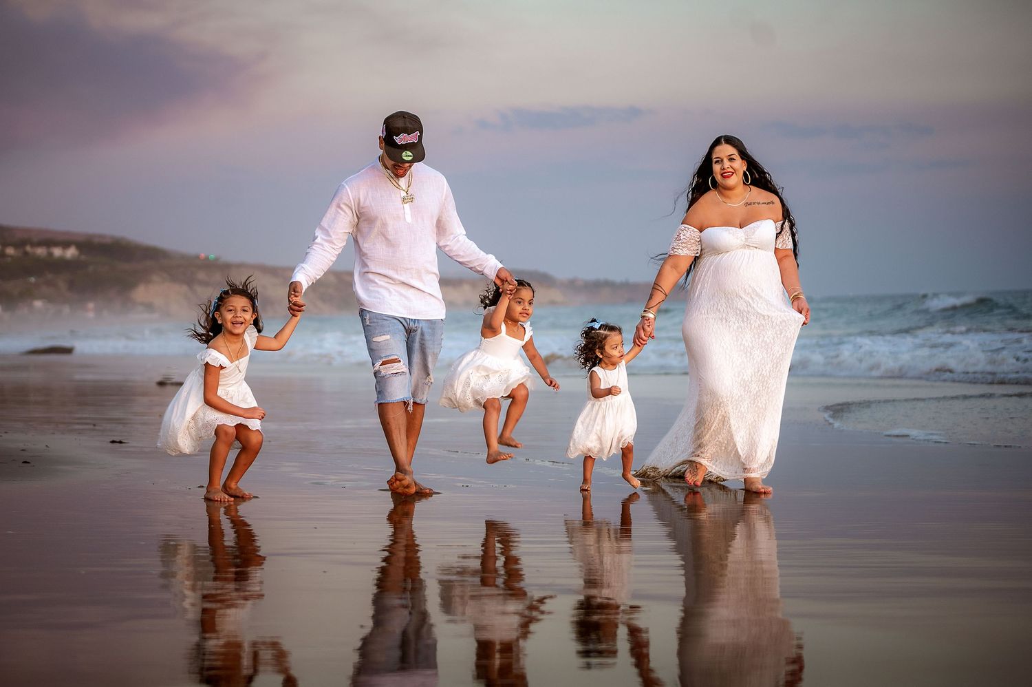 Family plays together at sunset on a sandy beach, wearing coordinated white outfits.