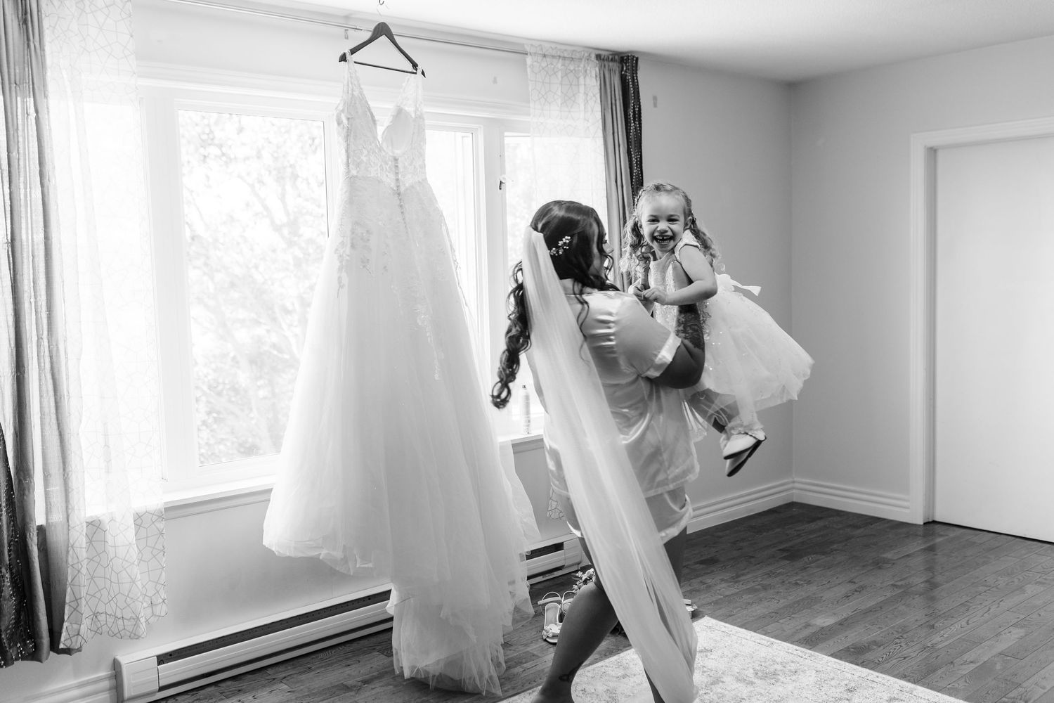 A white wedding dress hangs by a window while people prepare for a wedding ceremony in a black and white photo.