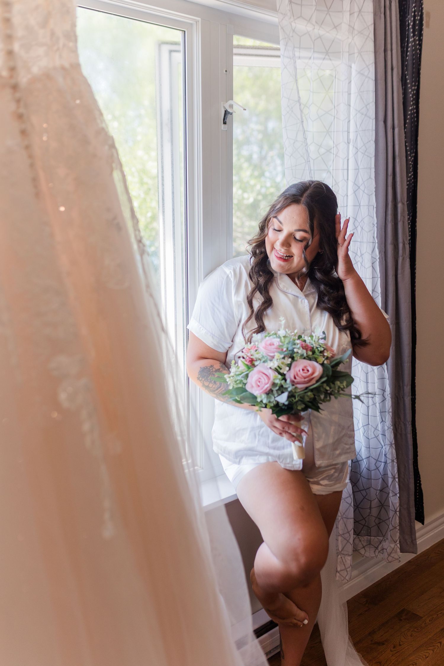 A romantic bridal boudoir photo shows a figure in white by a window holding a pink and blue floral bouquet.
