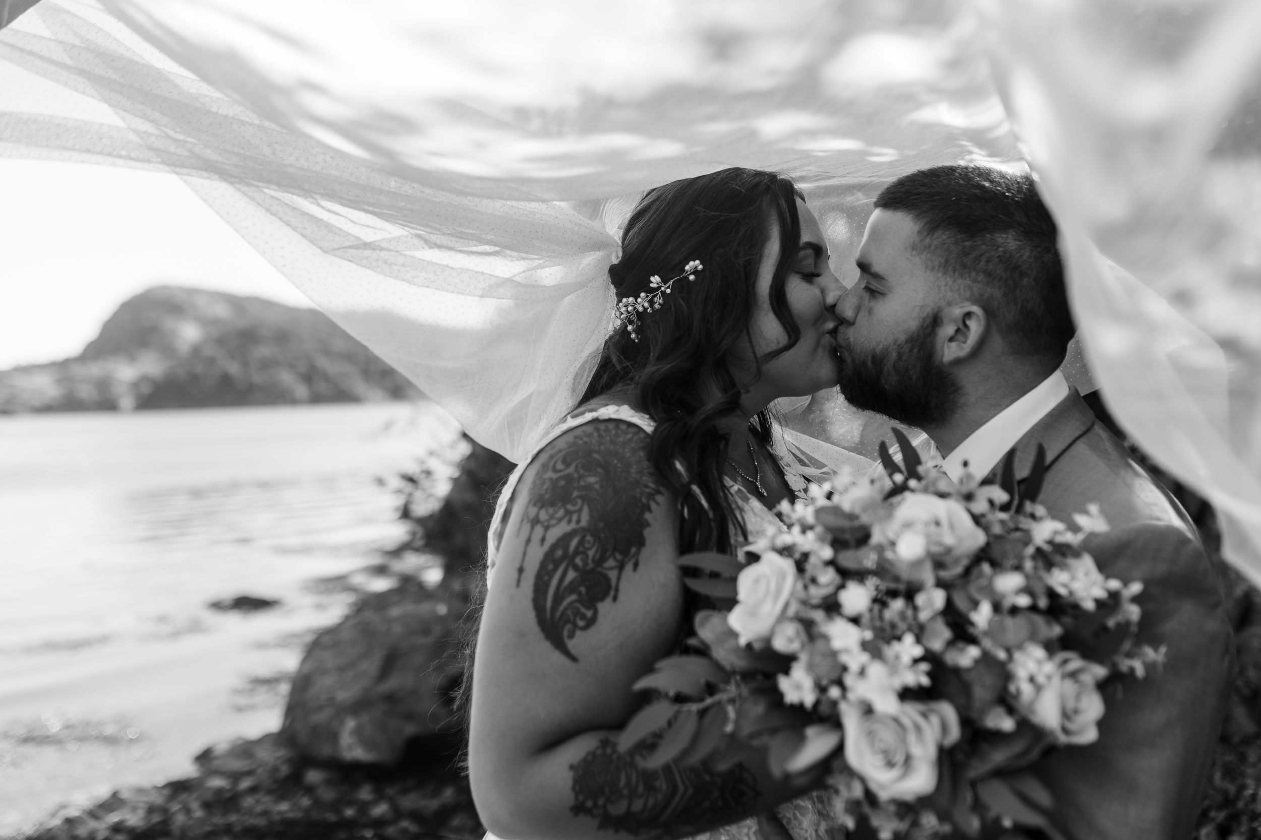 A romantic wedding kiss on a cliff with a mountain vista and floral bouquet in black and white.