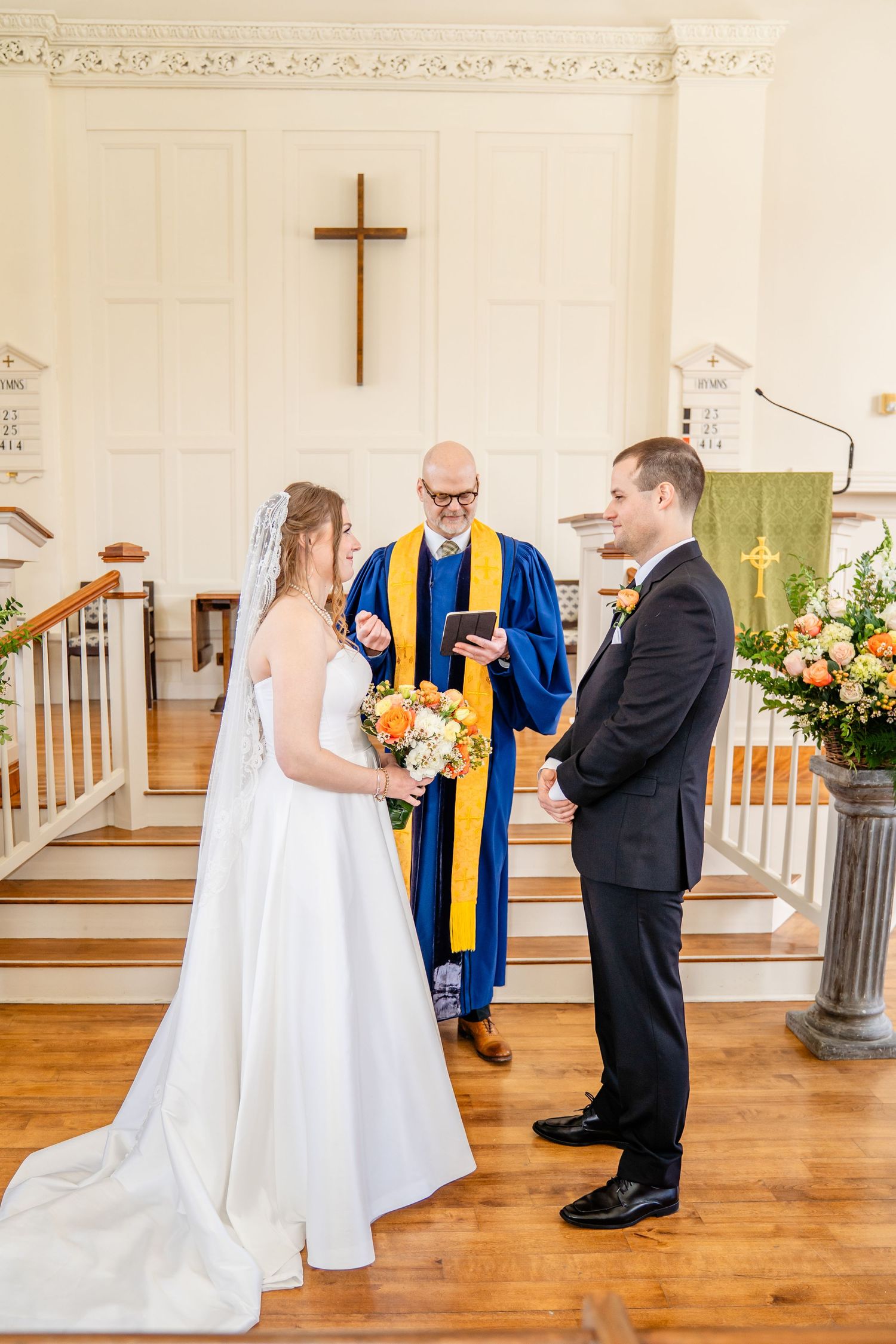 A wedding ceremony takes place inside a church with wooden floors and a cross on the wall during an intimate service.