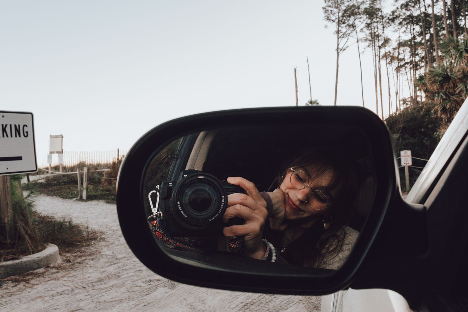 A DSLR camera and photographer are reflected in a car's side mirror on a rural road.