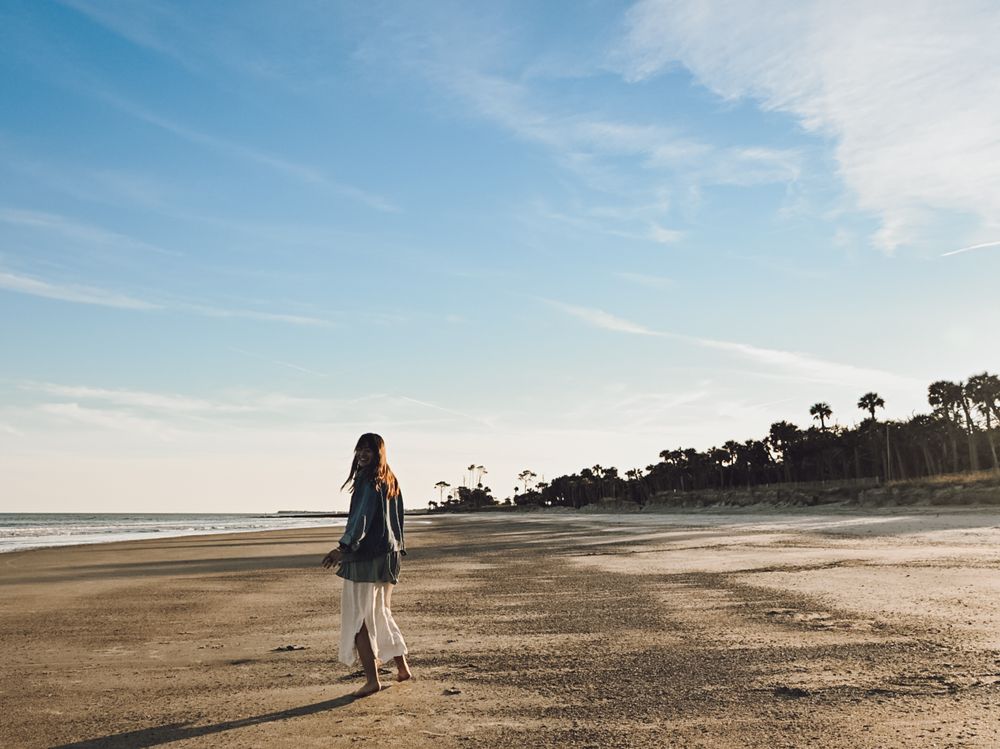 A person in a flowing dress walks alone on a serene sandy beach with palm trees and blue skies in the background.