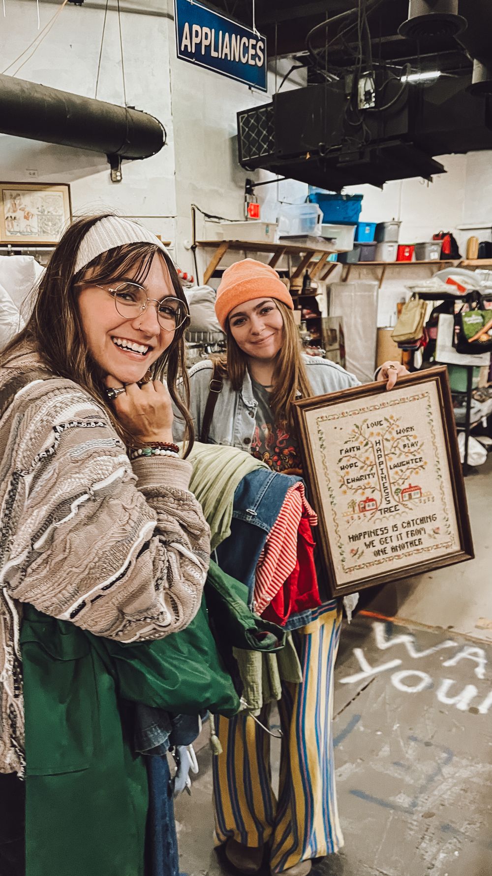 Two friends in thrift store smiling while holding vintage decor and clothes under an appliances sign.