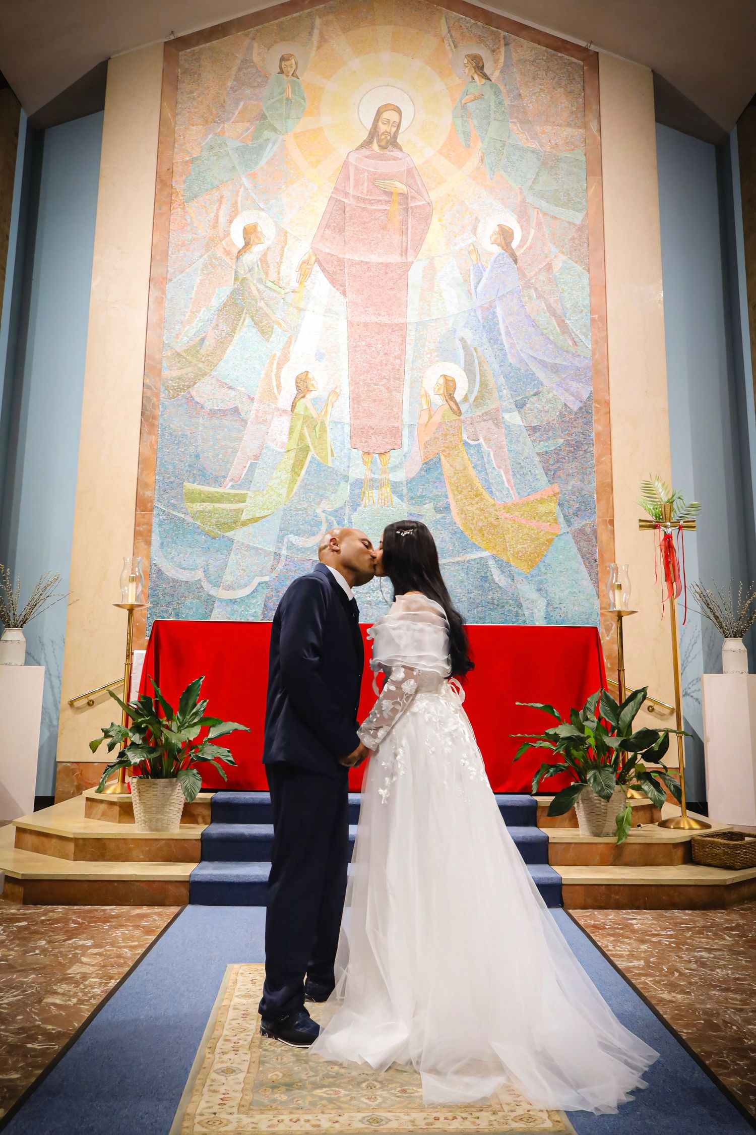 A wedding ceremony takes place inside a church with Jesus artwork and stained glass windows in the background.