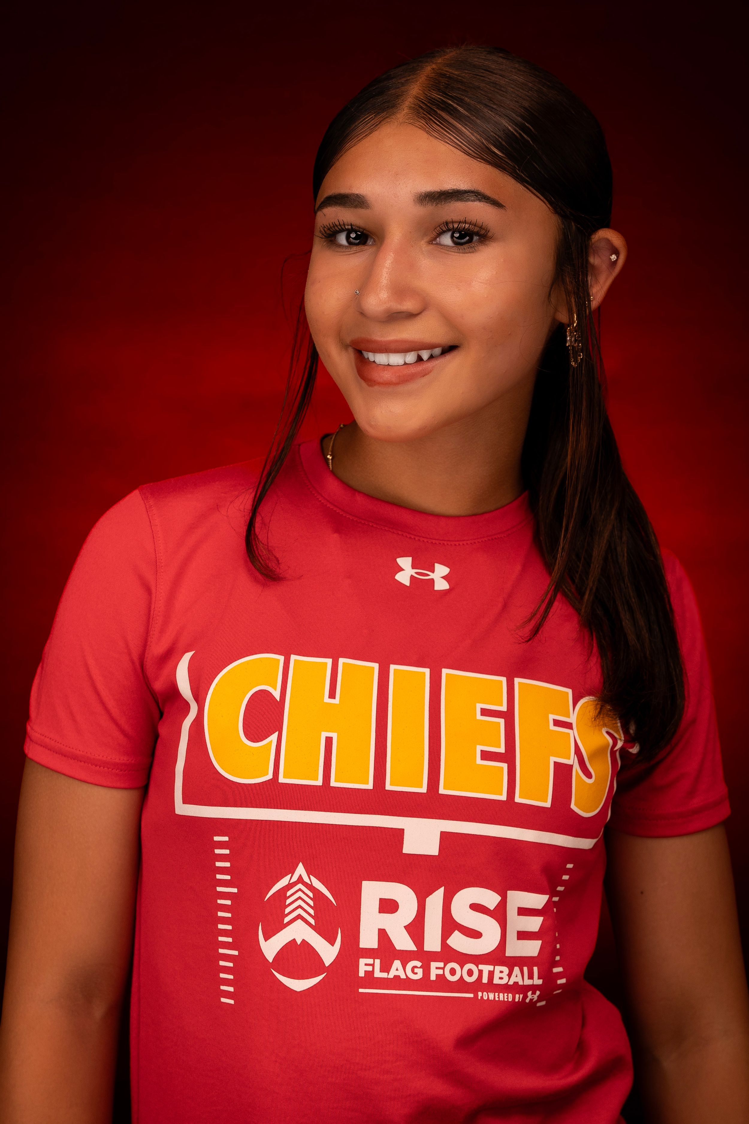 Young person in red Chiefs and Rise flag football shirt posing against dark red background.