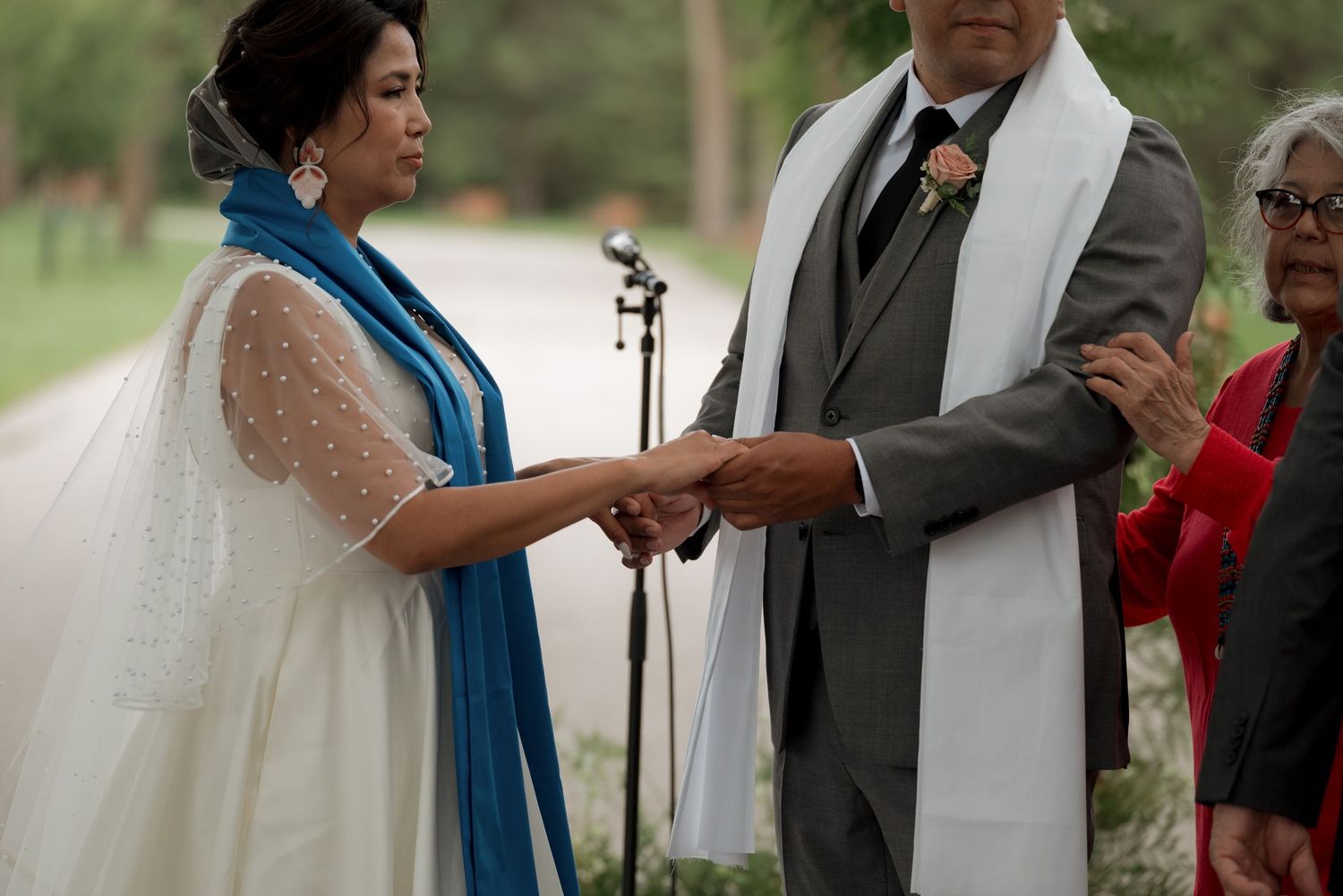Outdoor wedding ceremony with guests gathered under green trees.