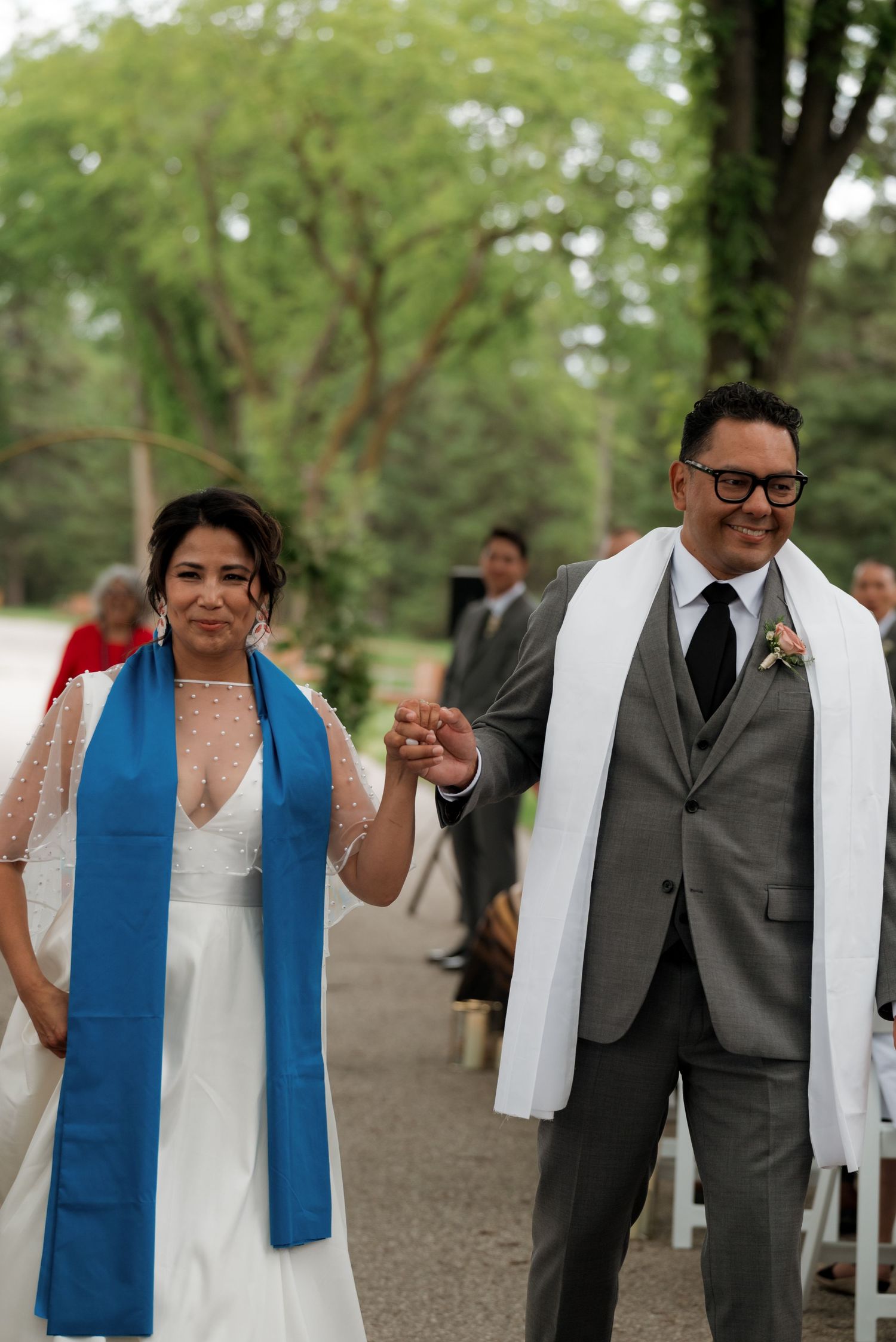 A couple celebrates at an outdoor wedding ceremony, wearing white and gray formal attire with a blue and white stoles.
