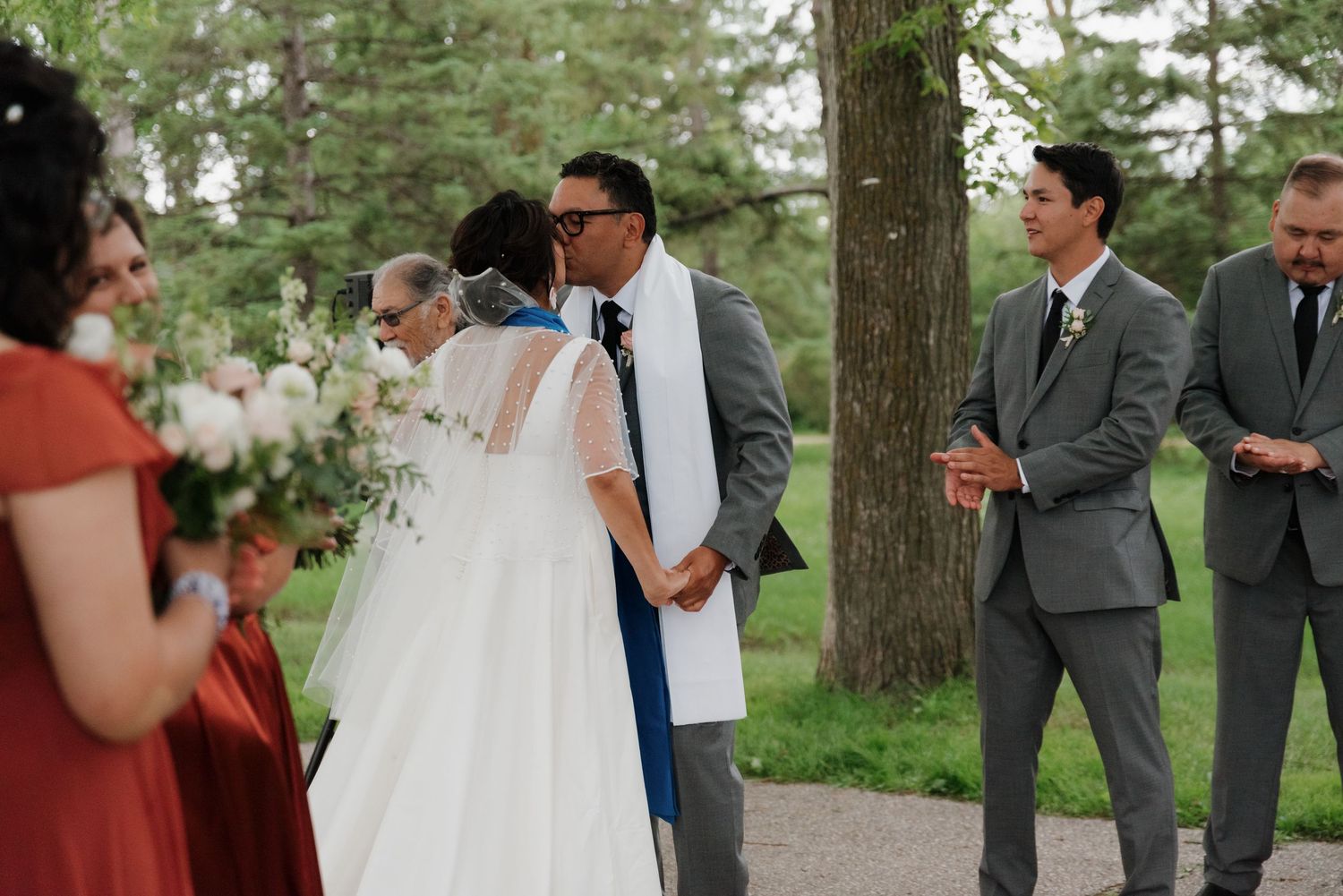 Outdoor wedding gathering with attendees in formal wear under mature trees.