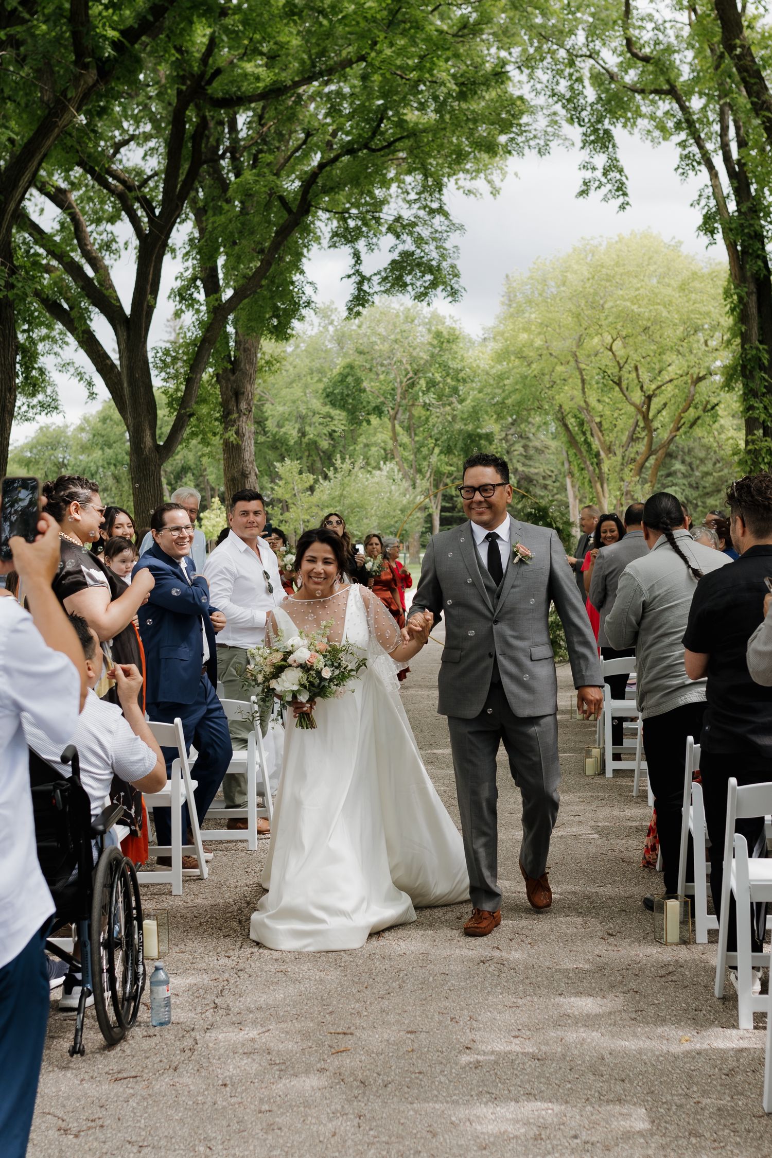 Outdoor wedding ceremony takes place under green trees with white chairs and guests gathered around.
