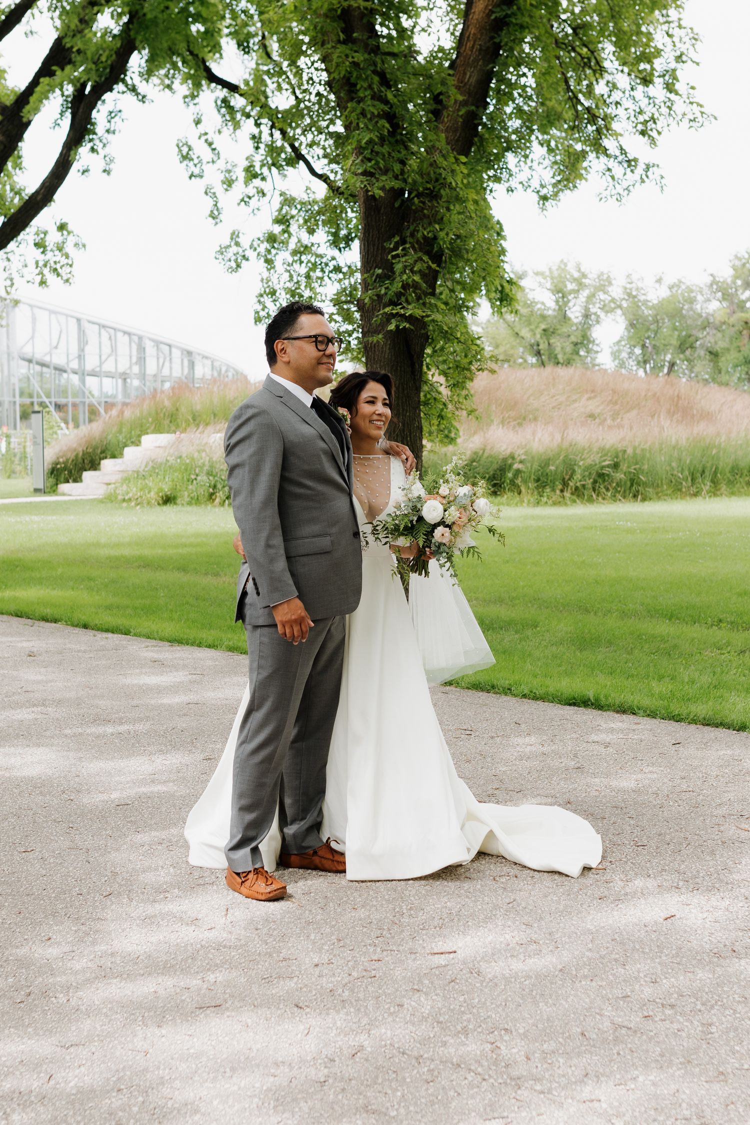 Wedding couple poses on gravel path surrounded by lush green scenery.