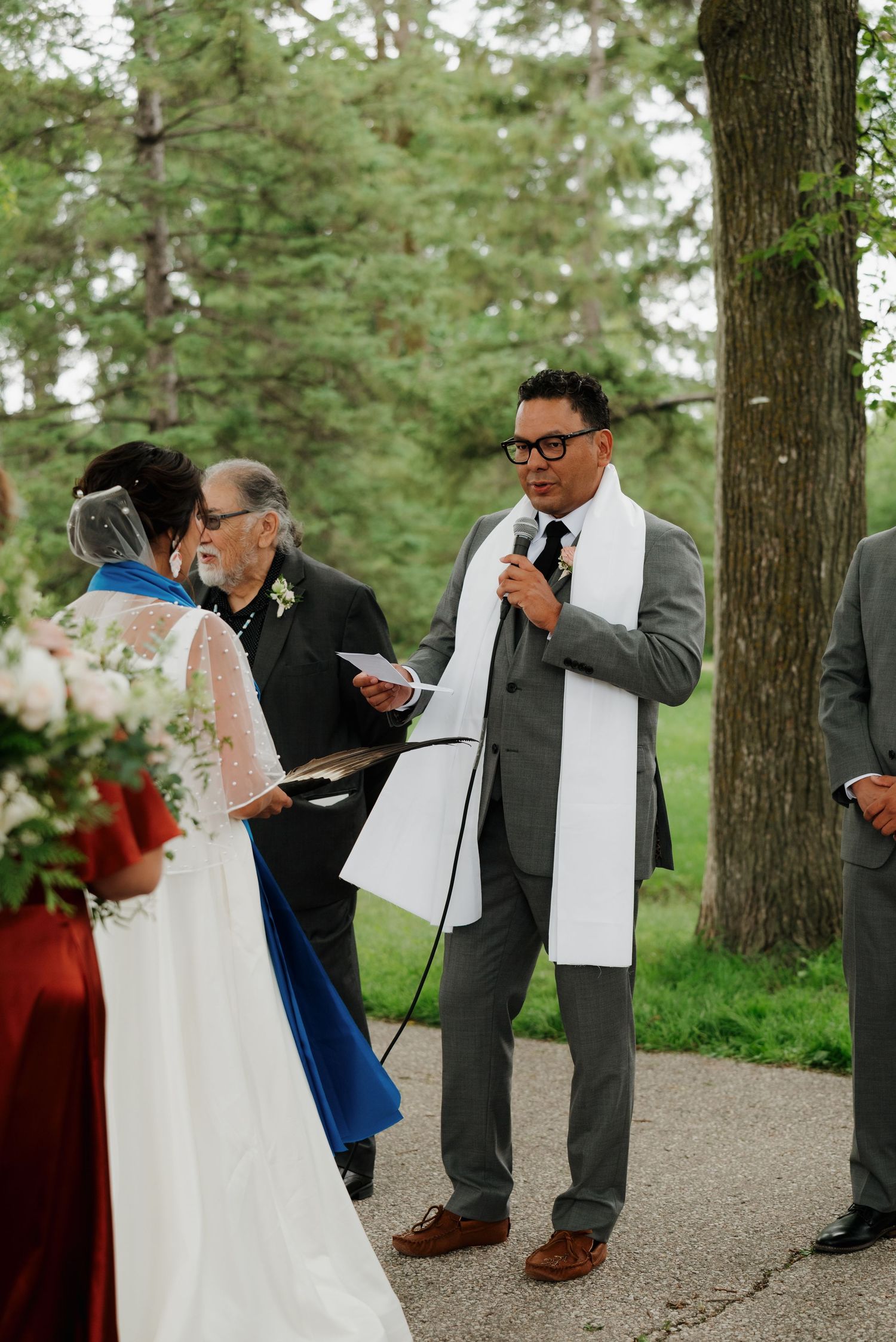 Wedding ceremony participants interacting during outdoor service under tall pine trees.