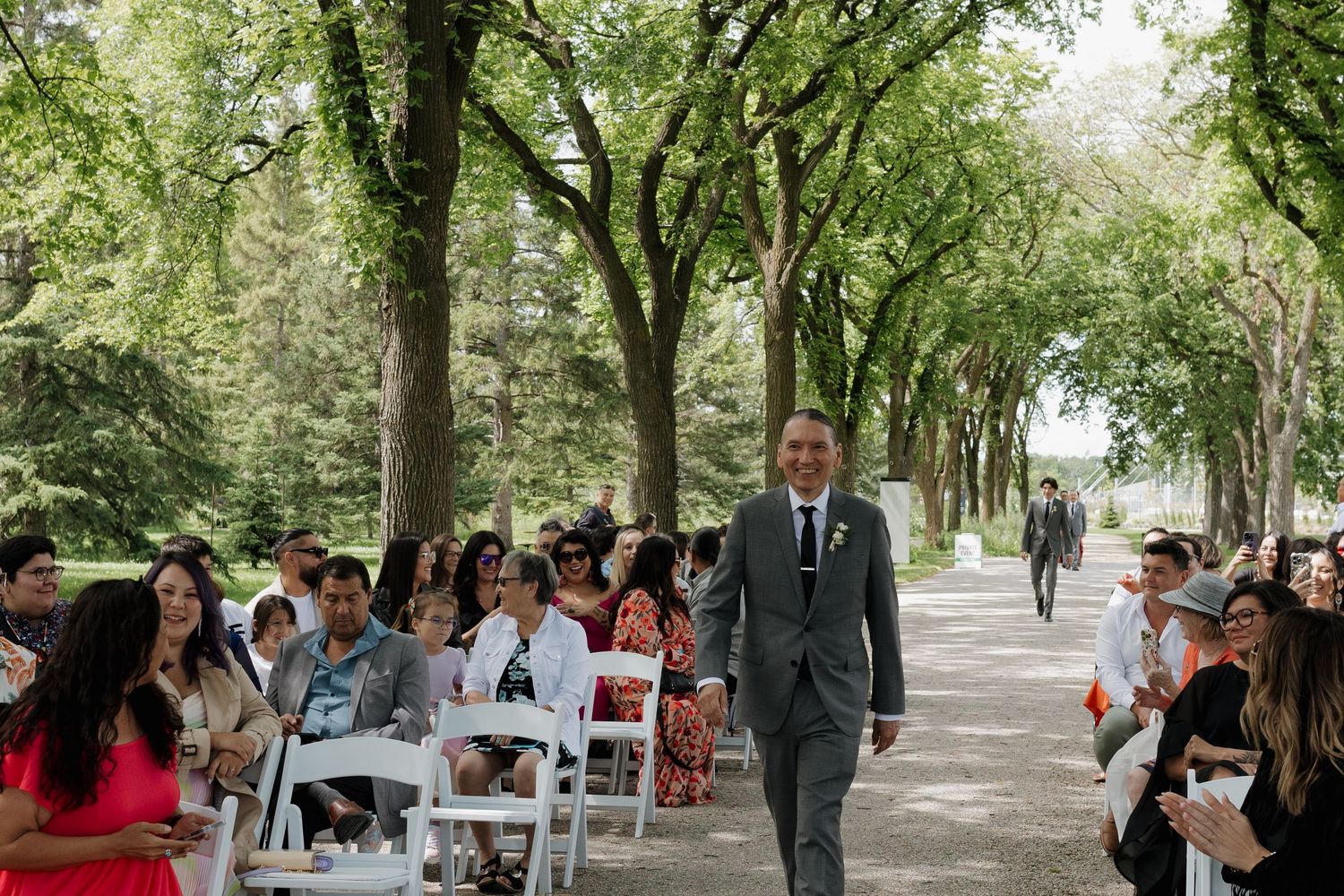 Outdoor wedding ceremony setup with rows of white chairs under a canopy of trees.