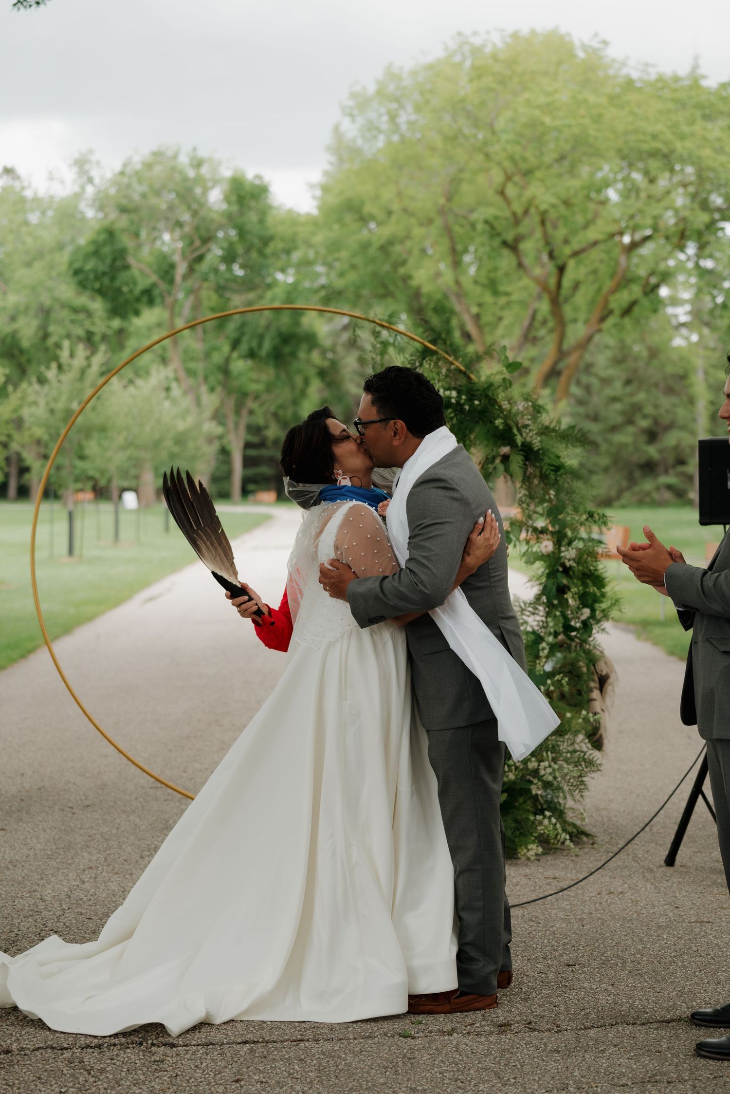 Romantic wedding couple poses with decorative floral hoop on path surrounded by trees.