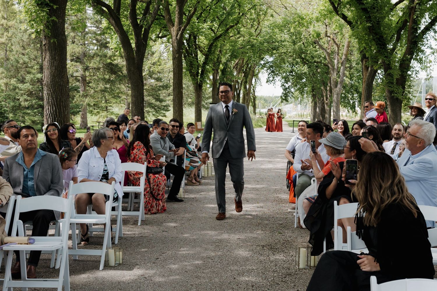 Guests gather under a canopy of trees at an outdoor event with white chairs and natural decor.