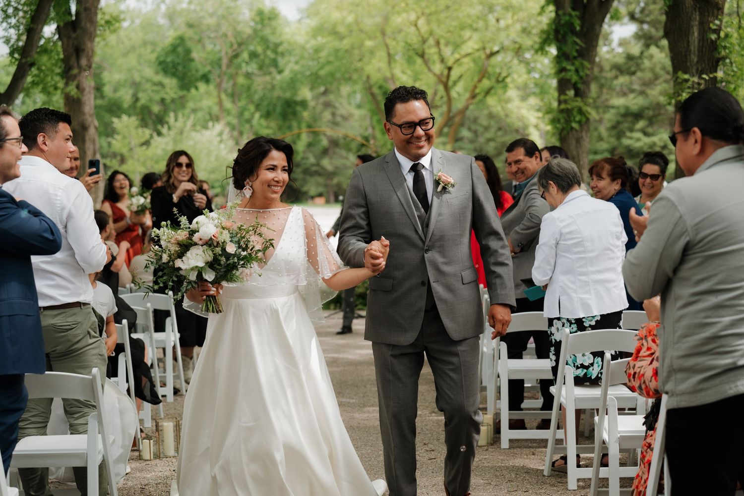 Wedding ceremony setup in a park with white chairs and guests gathering under tree cover.