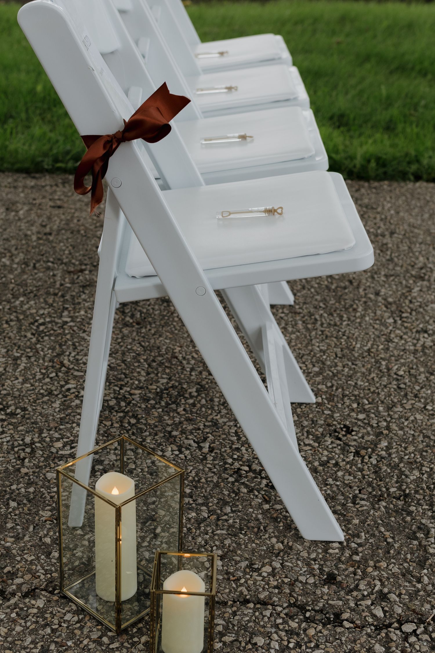 White wooden chairs and decorative lanterns arranged outdoors on a gravel surface for an elegant event setup.