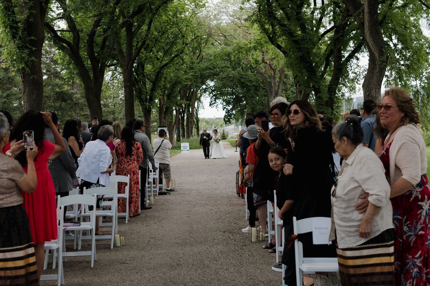 Outdoor wedding ceremony with white chairs lined up along a tree-covered path while guests gather before the event.