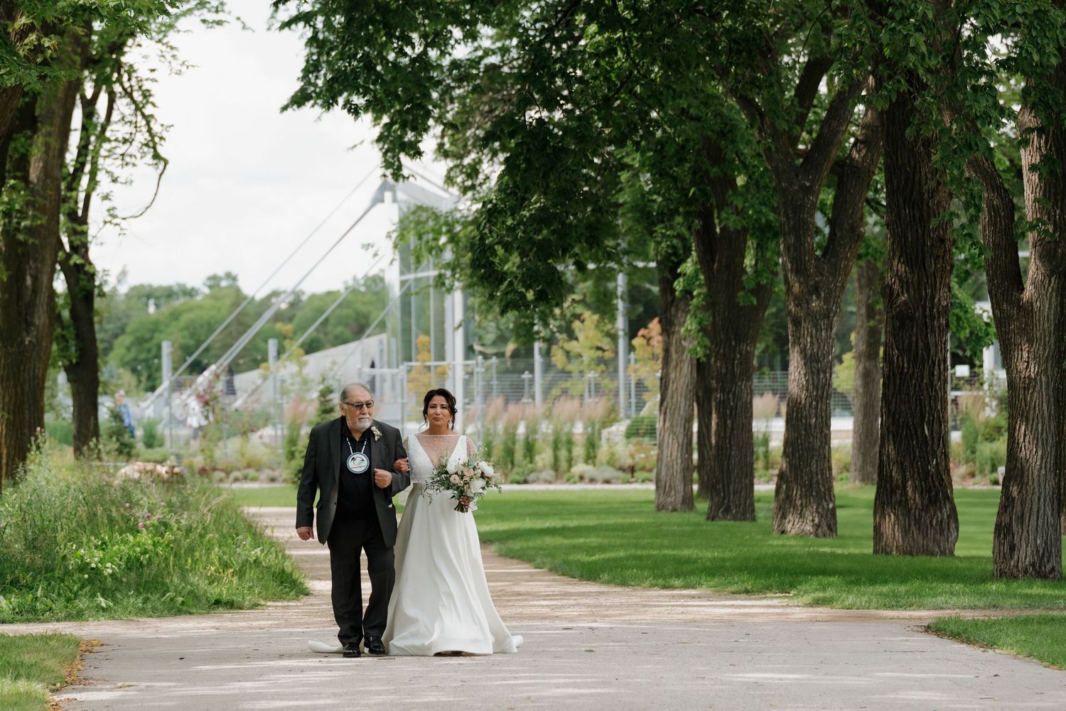 A romantic wedding ceremony takes place in a tree-lined outdoor path with a white gazebo in the background.
